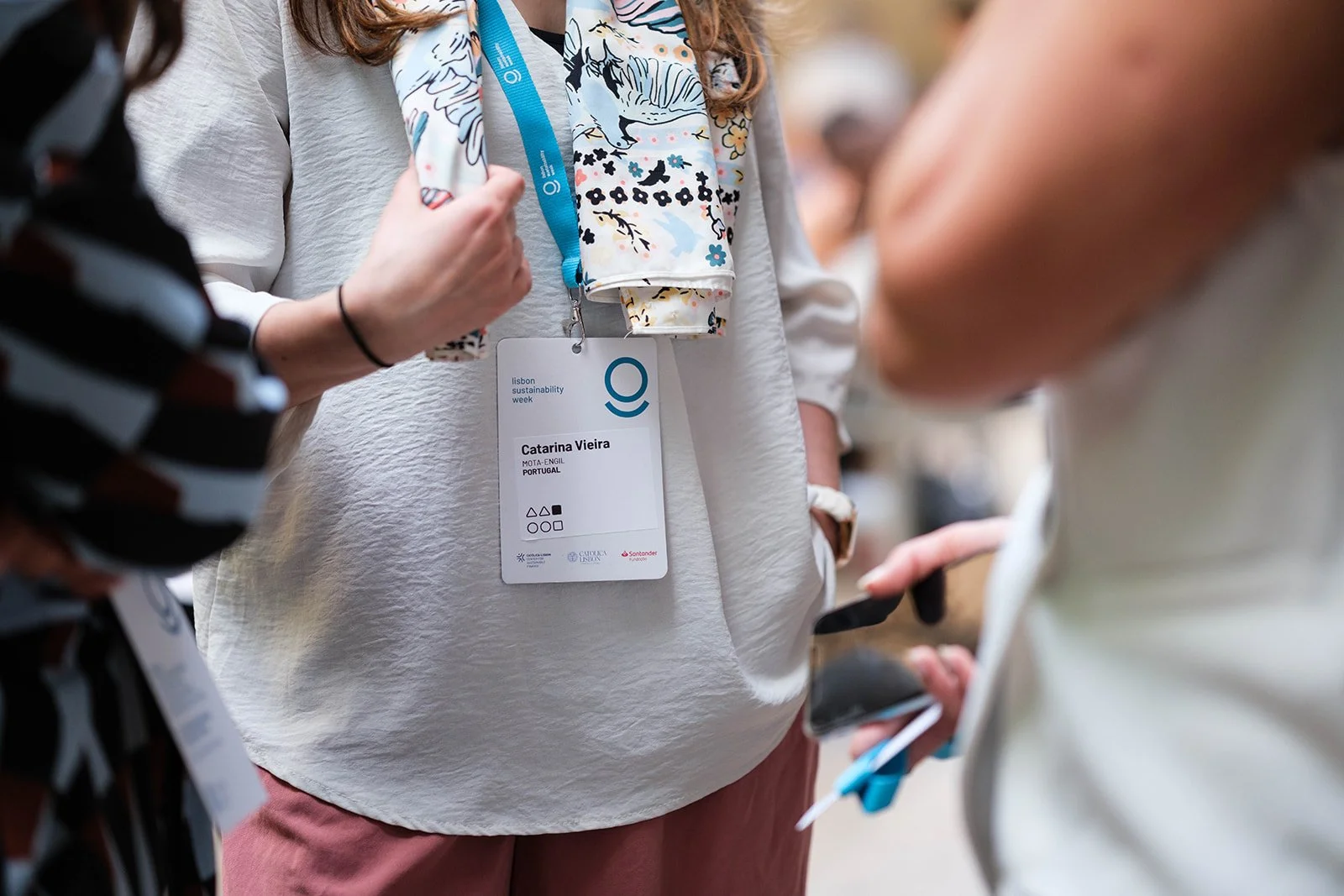 Close-up of a woman wearing a white top and pink pants with a lanyard and name tag. She is holding a colorful scarf. Other people are partially visible, some holding smartphones, at what seems to be a conference or event.