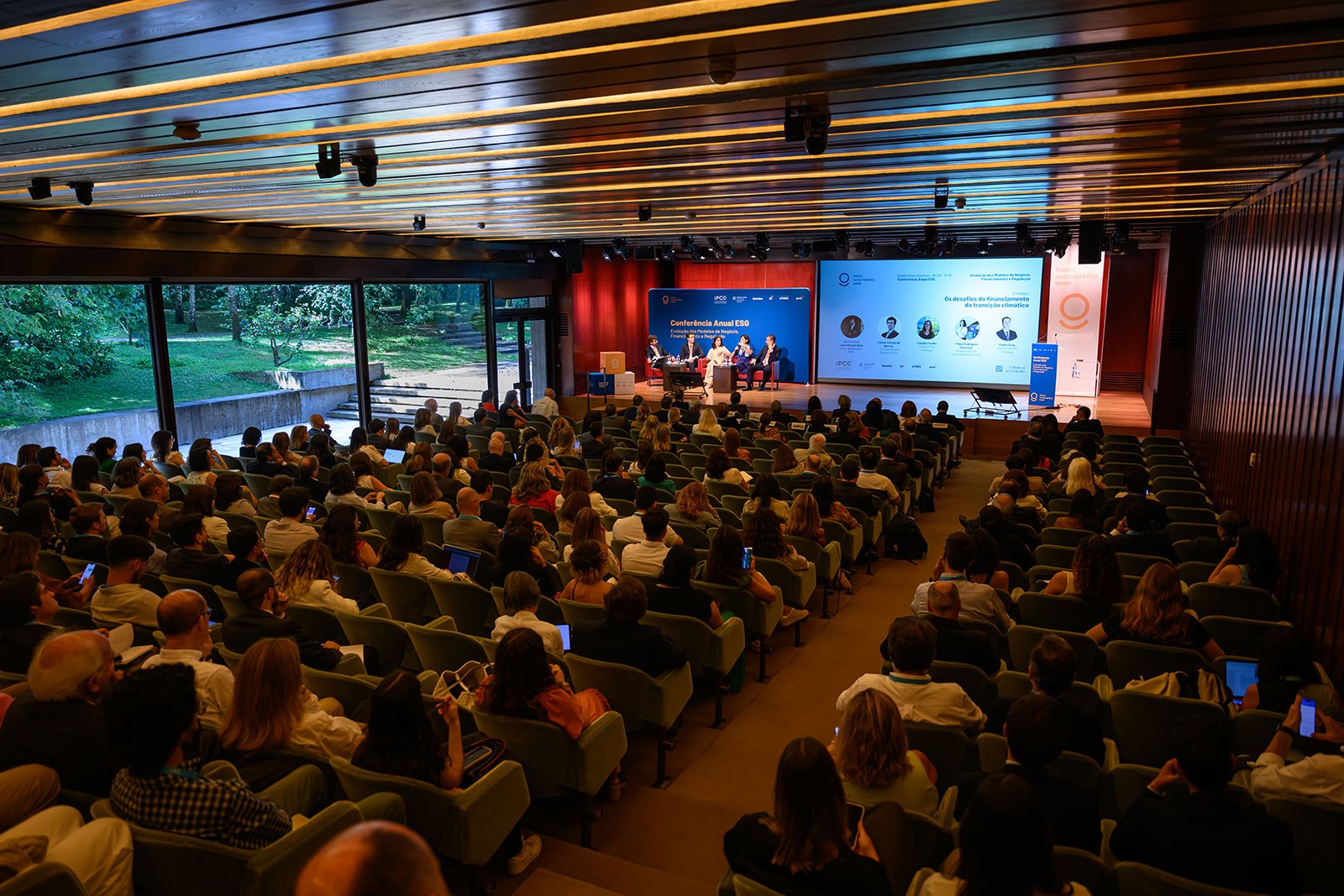 A large conference room filled with people attending a presentation on stage with panel speakers and screens displaying information.