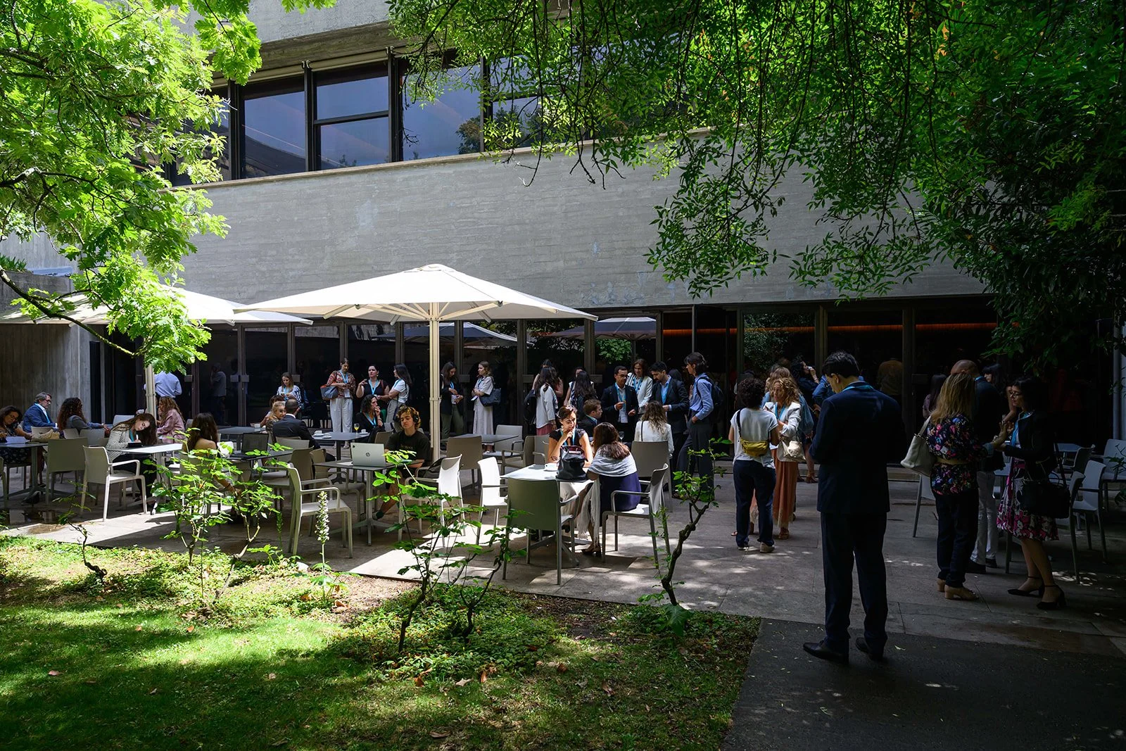 People socializing outside at a modern building with tables, chairs, large white umbrellas, and green trees.