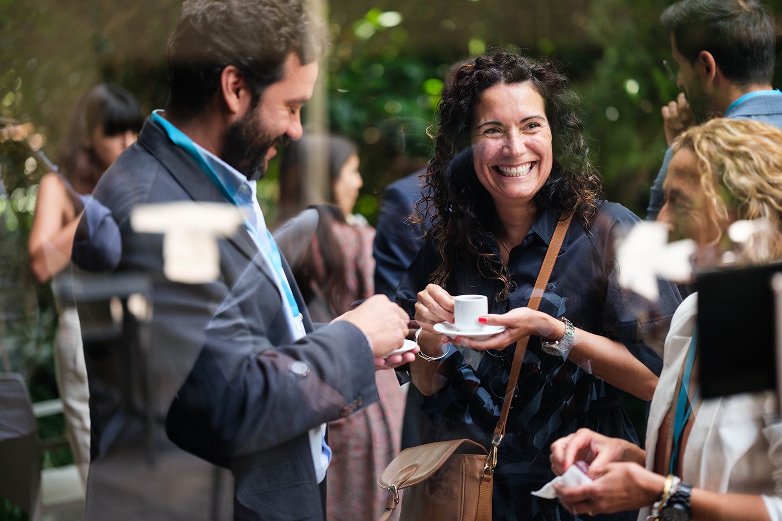 Group of people socializing outdoors, with a woman smiling and holding a small coffee cup, surrounded by others engaged in conversation.