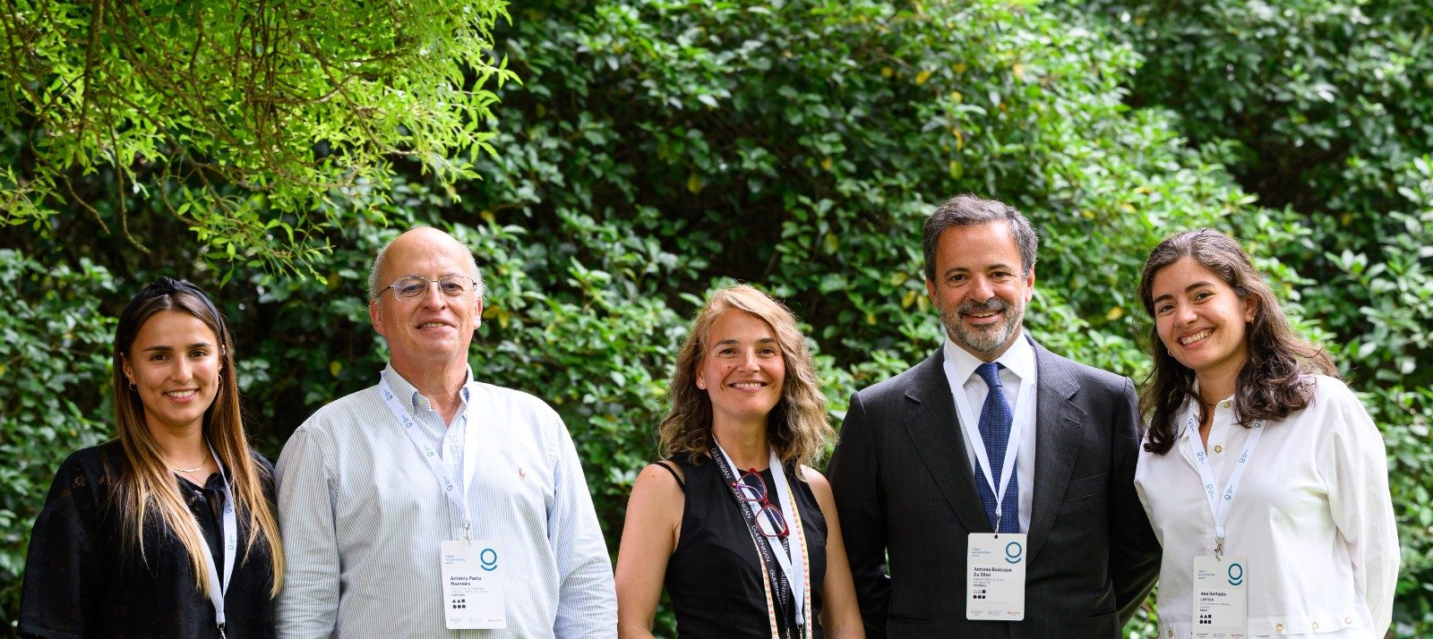 Group of five smiling people standing outdoors in front of green foliage.