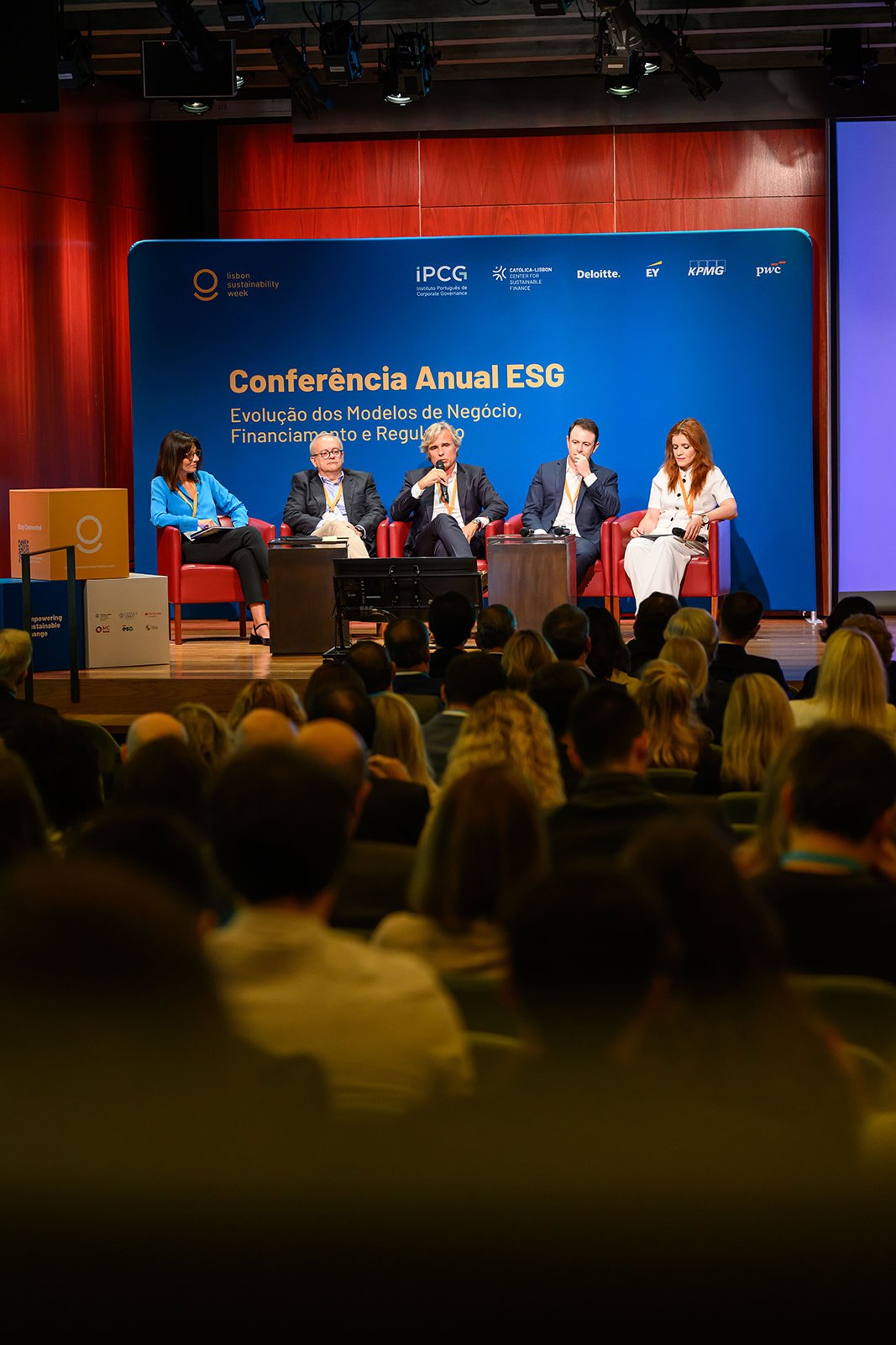 Panel discussion at a conference with five speakers seated on stage in front of an audience. The blue backdrop displays text referencing an annual ESG conference and various corporate sponsors.