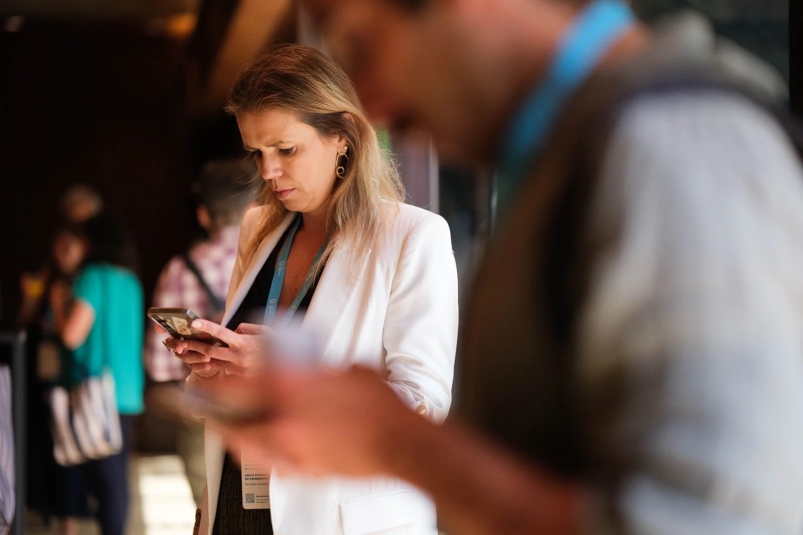 A woman in a white blazer looks at her phone with a focused expression while a person in the foreground also looks at a phone. Several people are blurred in the background.