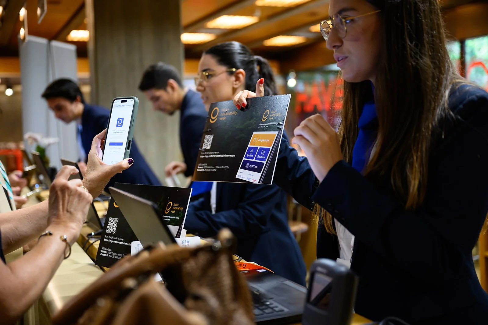 People at a conference counter, one person is holding a brochure and another is using a smartphone to scan a QR code.