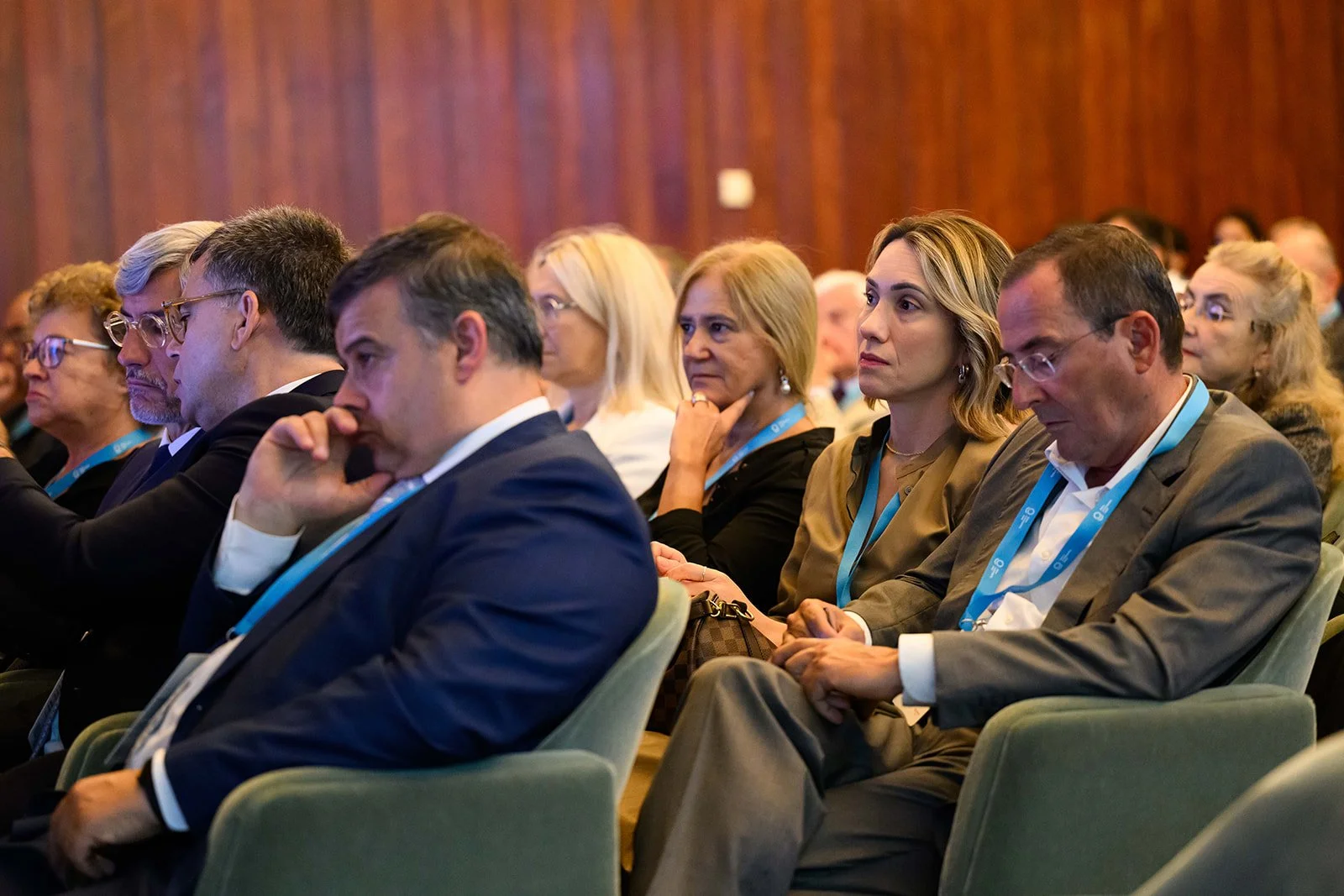 Conference attendees sitting in a row, listening attentively, wearing business attire and conference badges.