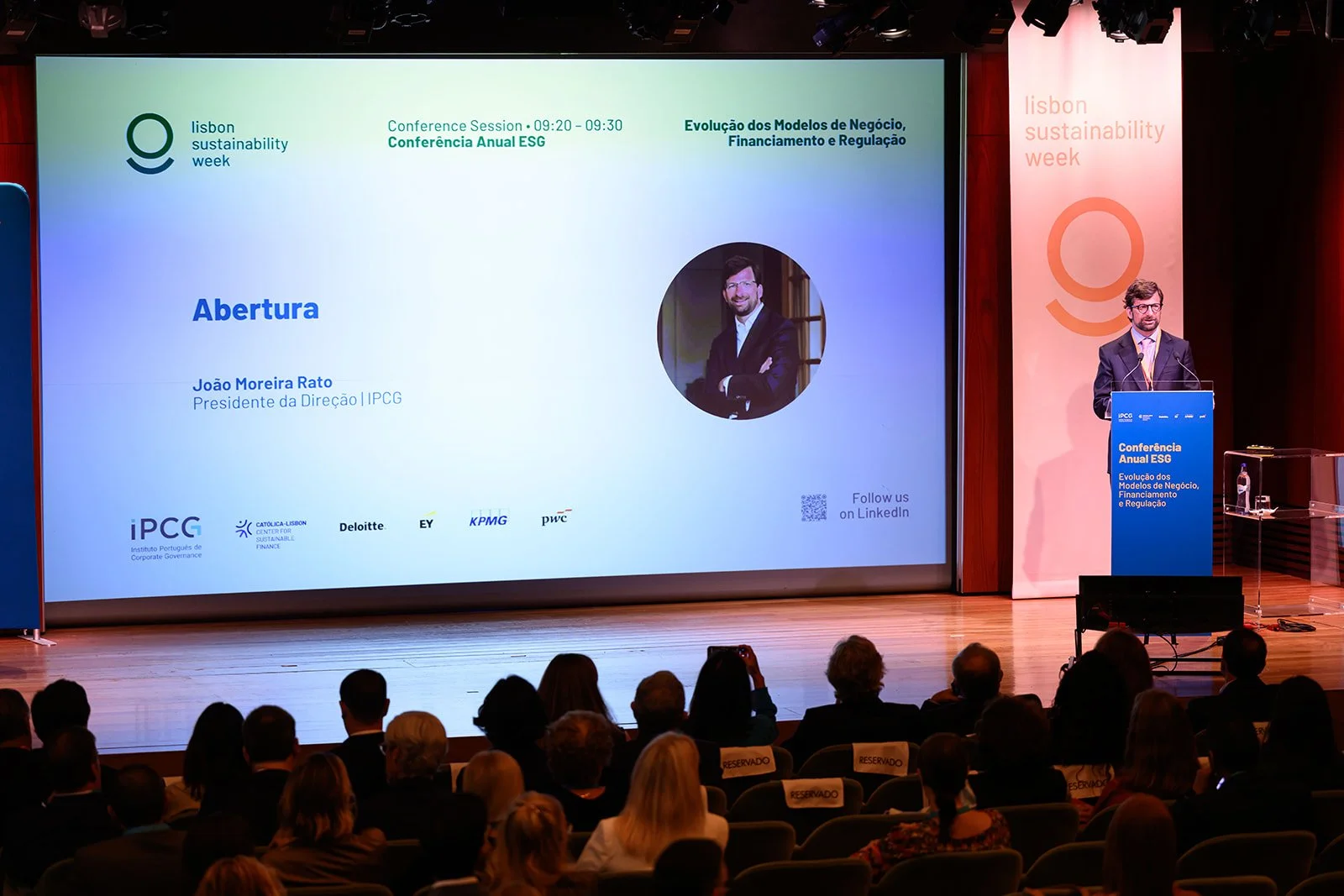A man in a suit stands at a blue podium on stage speaking at the Lisbon Sustainability Week conference. Behind him, a large screen displays a presentation slide with a photo of João Moreira Rato and event information. The audience is seated and watch
