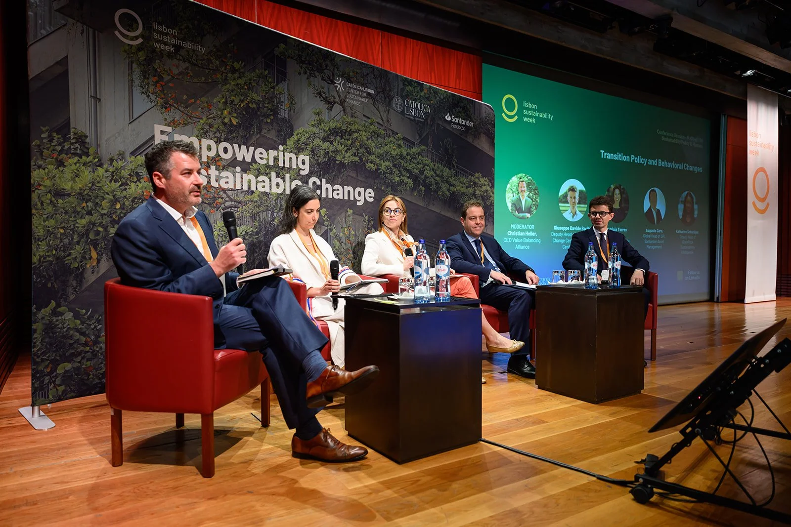 Panel discussion at Lisbon sustainability week with five people seated on stage, two women and three men, engaged in conversation. A large screen in the background displays the event title and speakers. The stage has a wooden floor and is decorated w