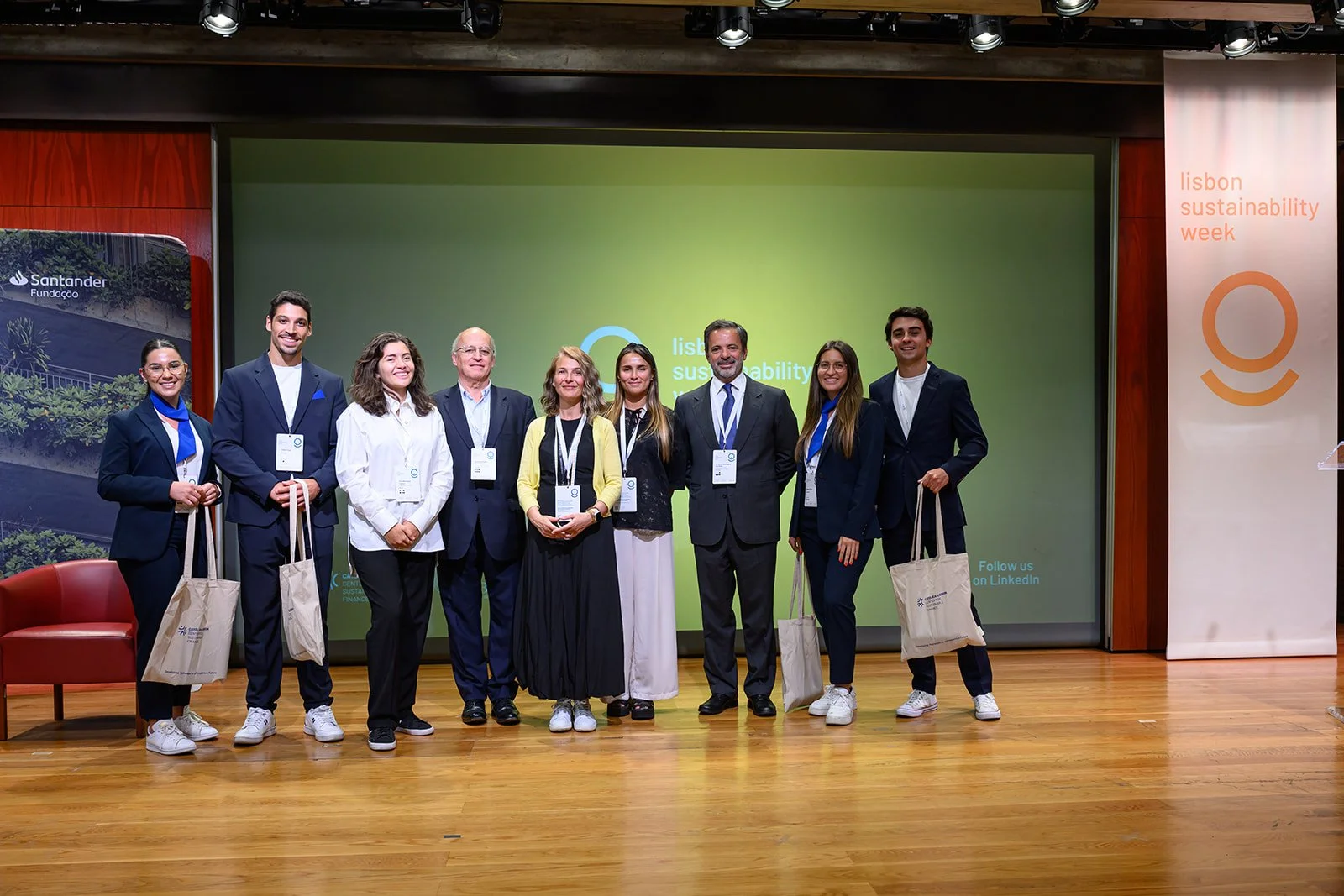 Group of nine diverse people standing on stage at Lisbon Sustainability Week, smiling, wearing name badges, some holding tote bags, in front of a green screen with event branding.