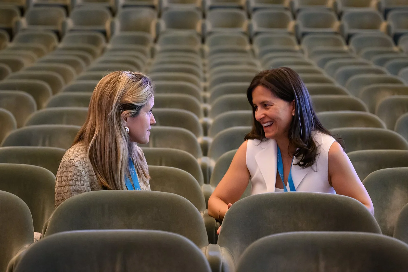 Two women seated in an empty auditorium, engaged in a friendly conversation, smiling at each other.