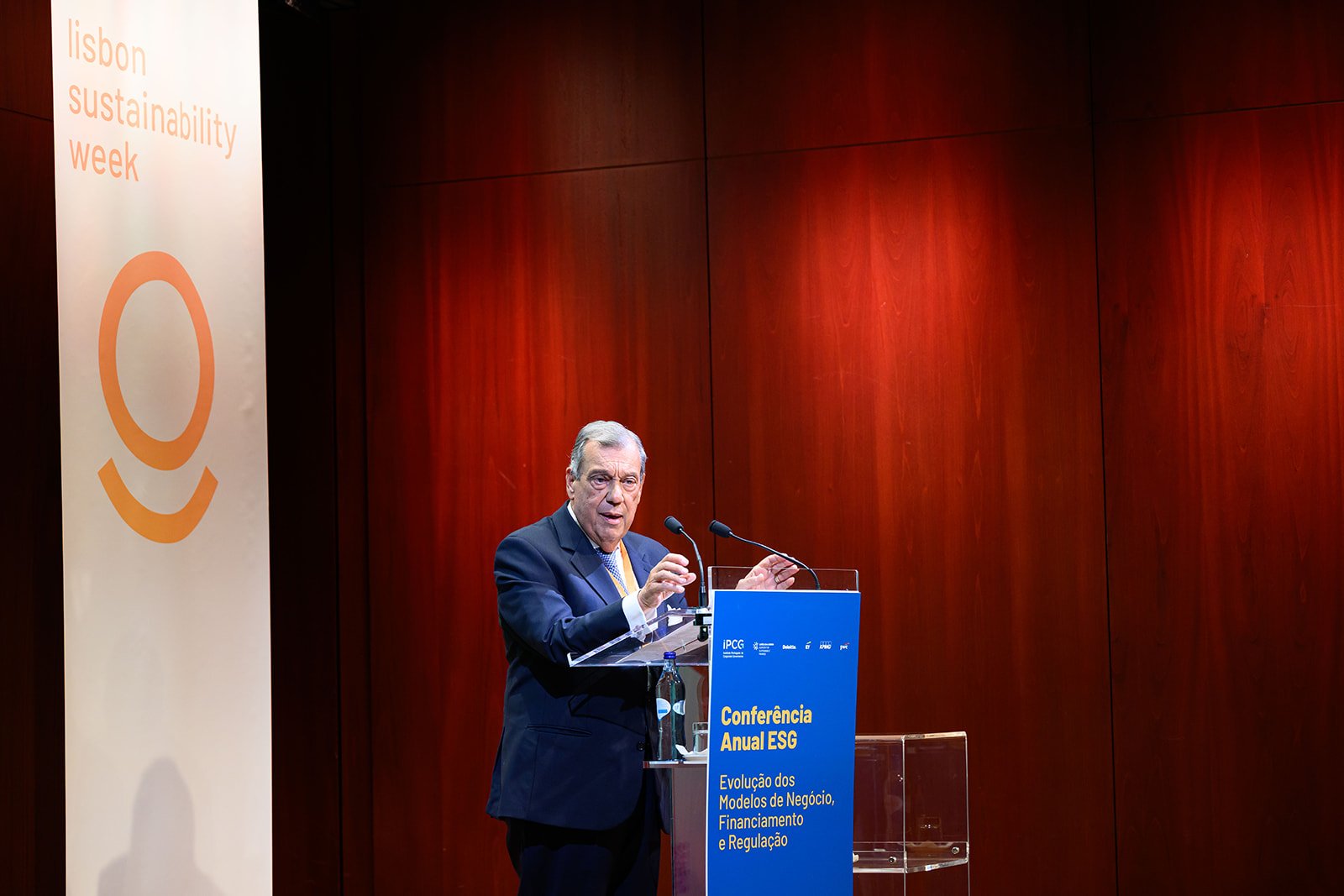 A man in a suit giving a speech at a podium during a conference about ESG, with a large vertical banner on the left that says 'Lisbon sustainability week' and a blue sign on the podium that reads 'Conferência Anual ESG, Evolução dos Modelos de Negóci