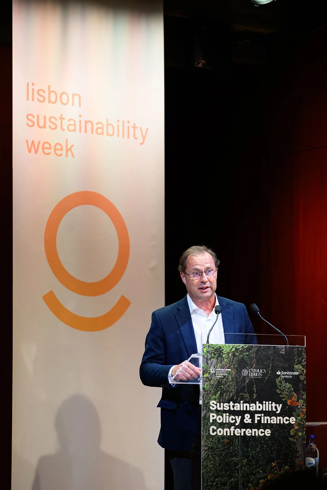 A man in formal attire giving a speech at a conference with a sign behind him that reads "lisbon sustainability week". The podium has a sign that says "Sustainability Policy & Finance Conference".