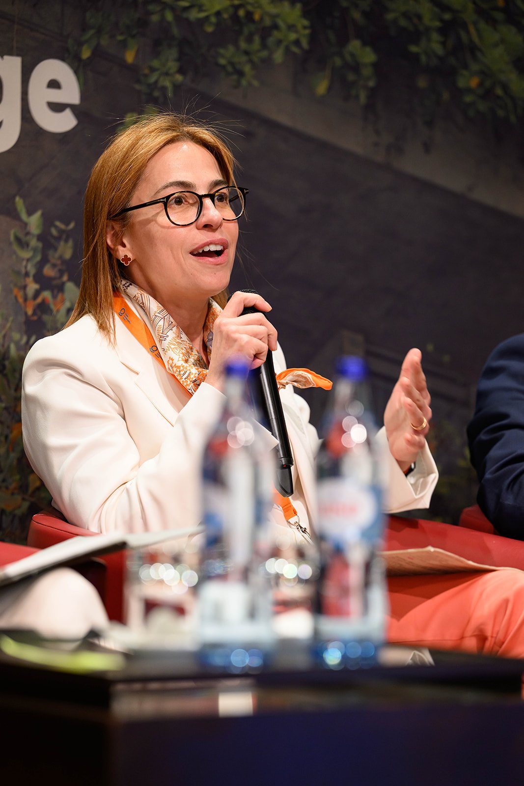 A woman with glasses, wearing a white blazer and orange scarf, speaking into a microphone at a panel discussion or conference. There are water bottles and papers on the table in front of her.