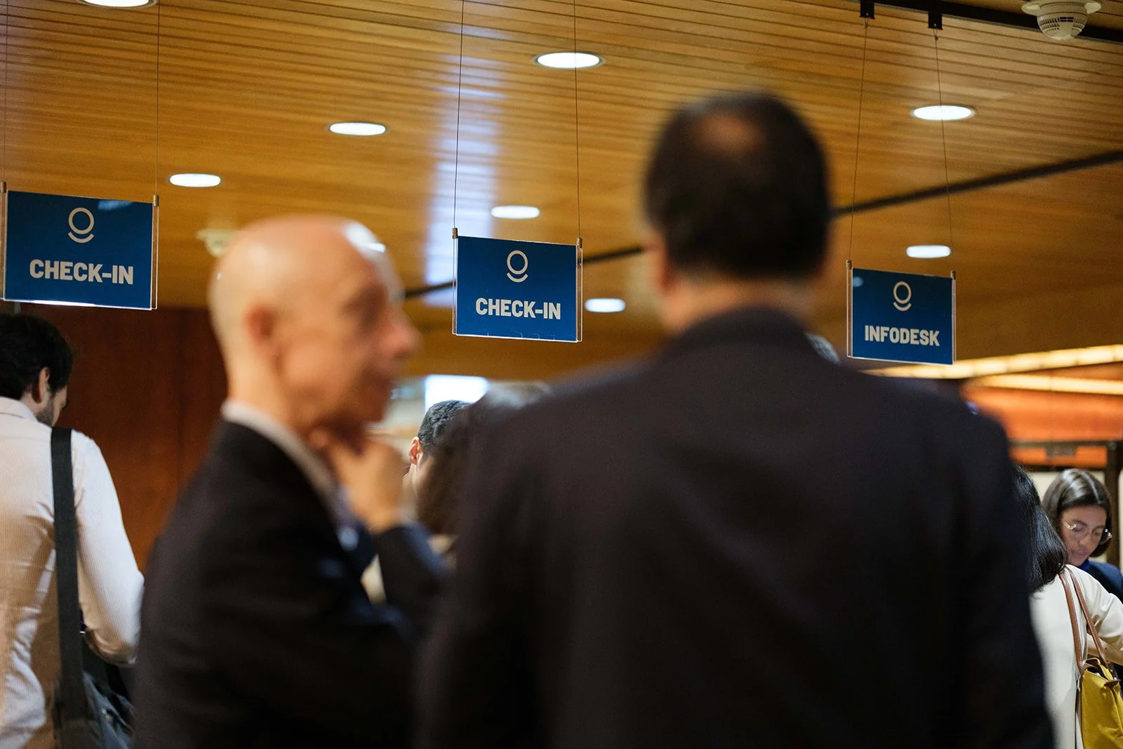People standing at check-in and infodesk counters in an indoor setting with wooden ceiling panels, illuminated by round ceiling lights.