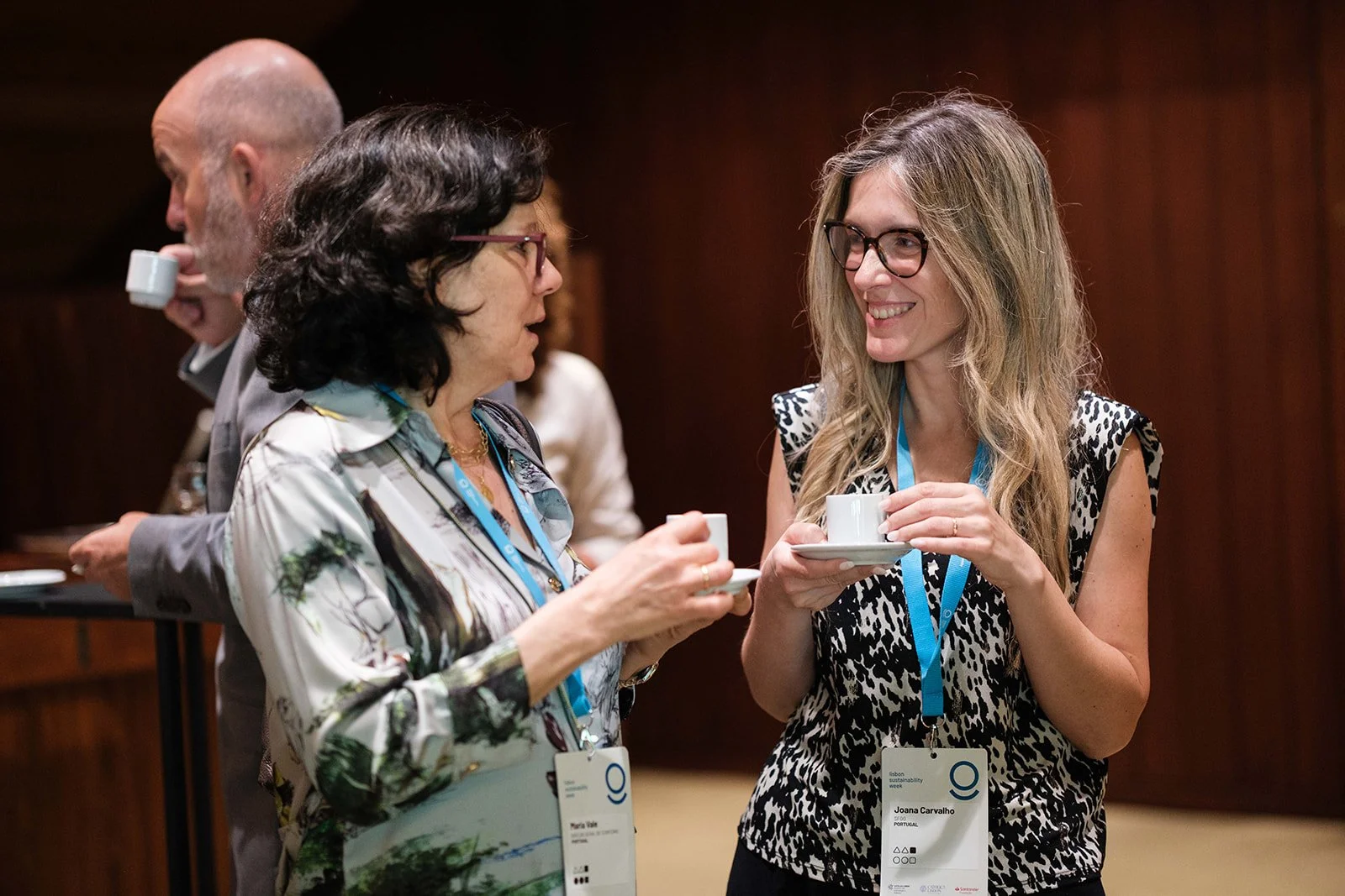 Two women and a man at a professional event, engaged in conversation, holding coffee cups. The women are smiling and wearing name tags on blue lanyards. The setting appears to be an indoor conference or networking event.