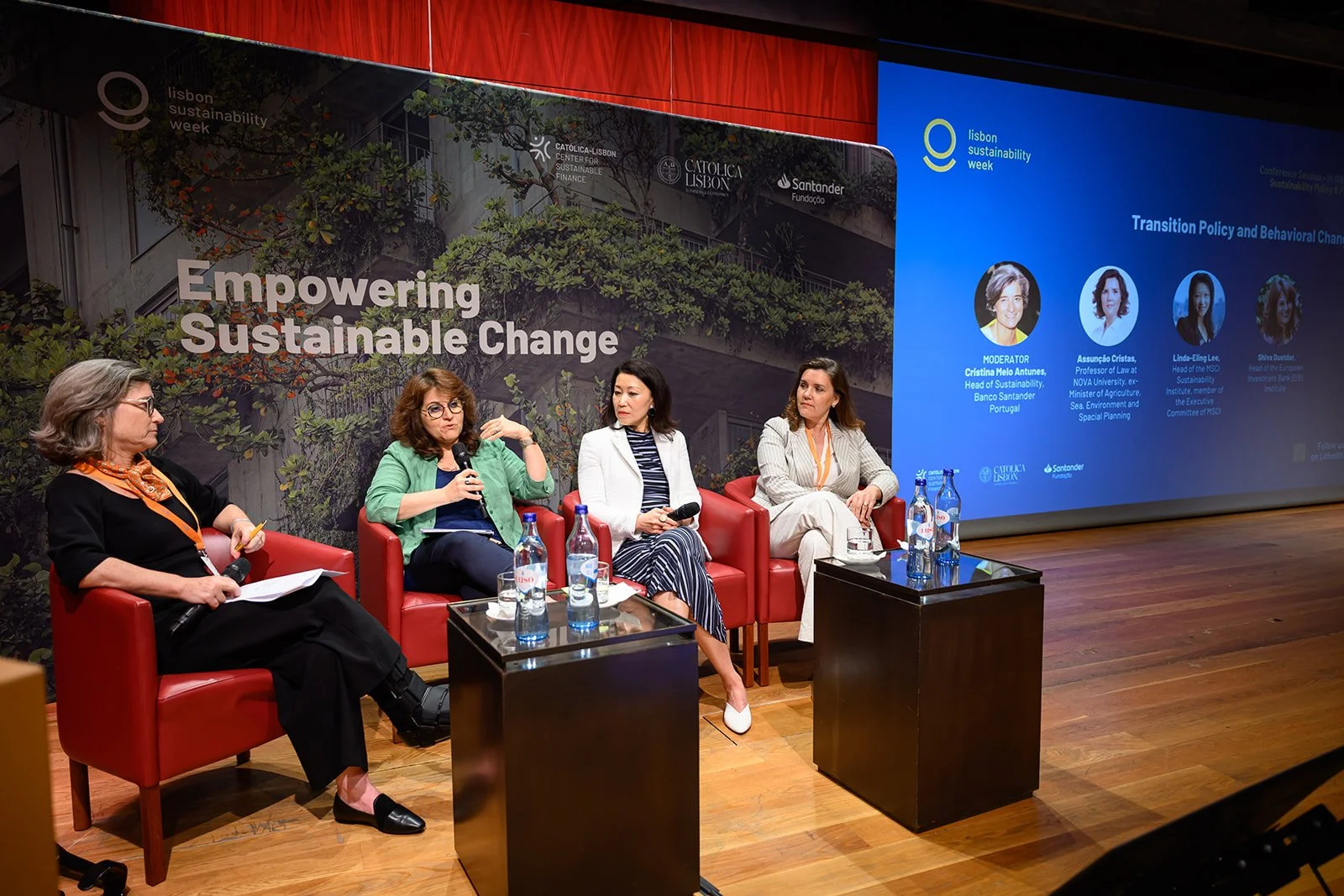 Four women seated on red chairs participating in a panel discussion at Lisbon Sustainability Week, with a backdrop displaying the event slogan and a large screen showing panel details.