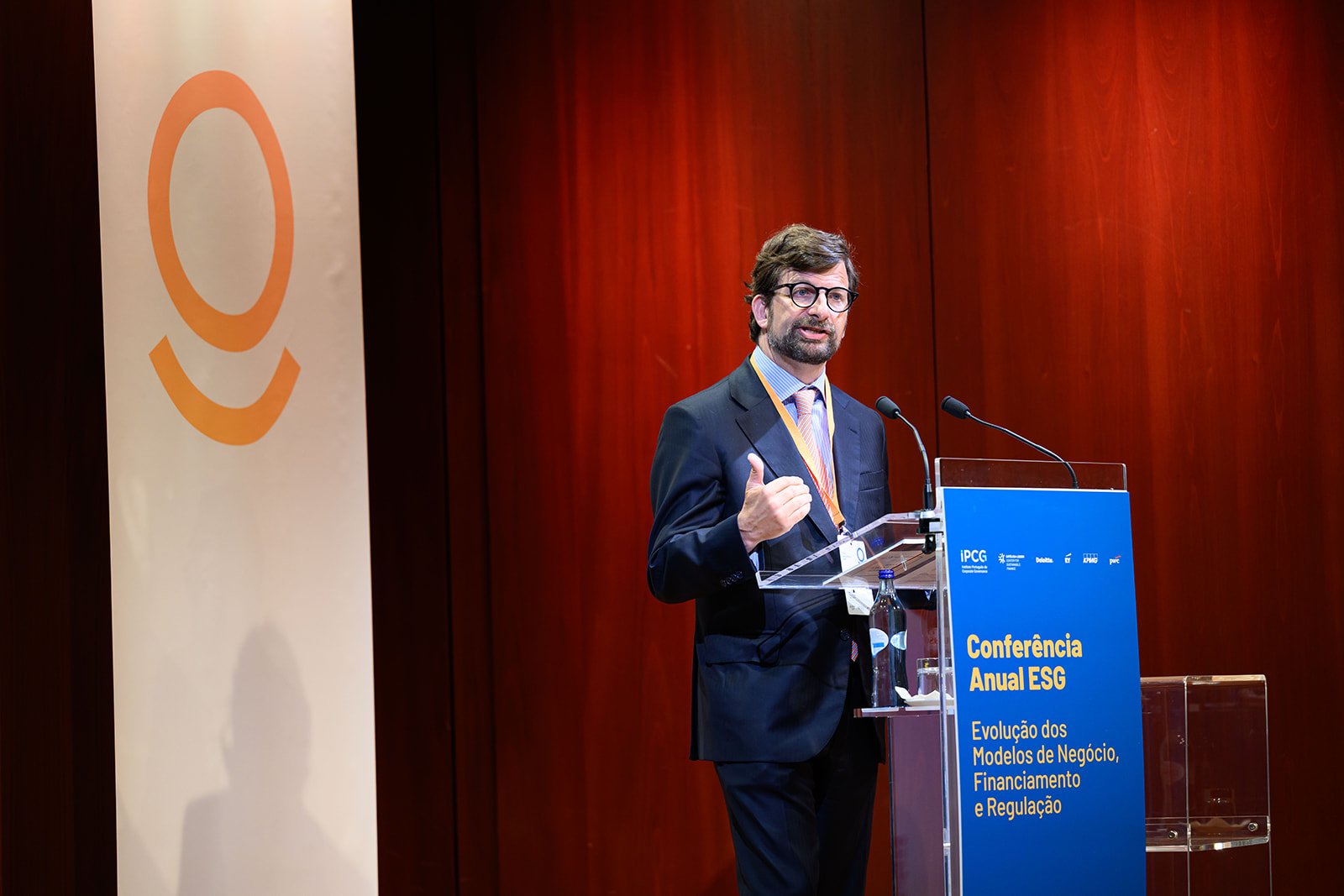 A man in a suit and glasses standing at a podium giving a presentation at a conference, with a blue and yellow sign that reads "Conferência Anual ESG" and "Evolução dos Modelos de Negócio, Financiamento e Regulação."