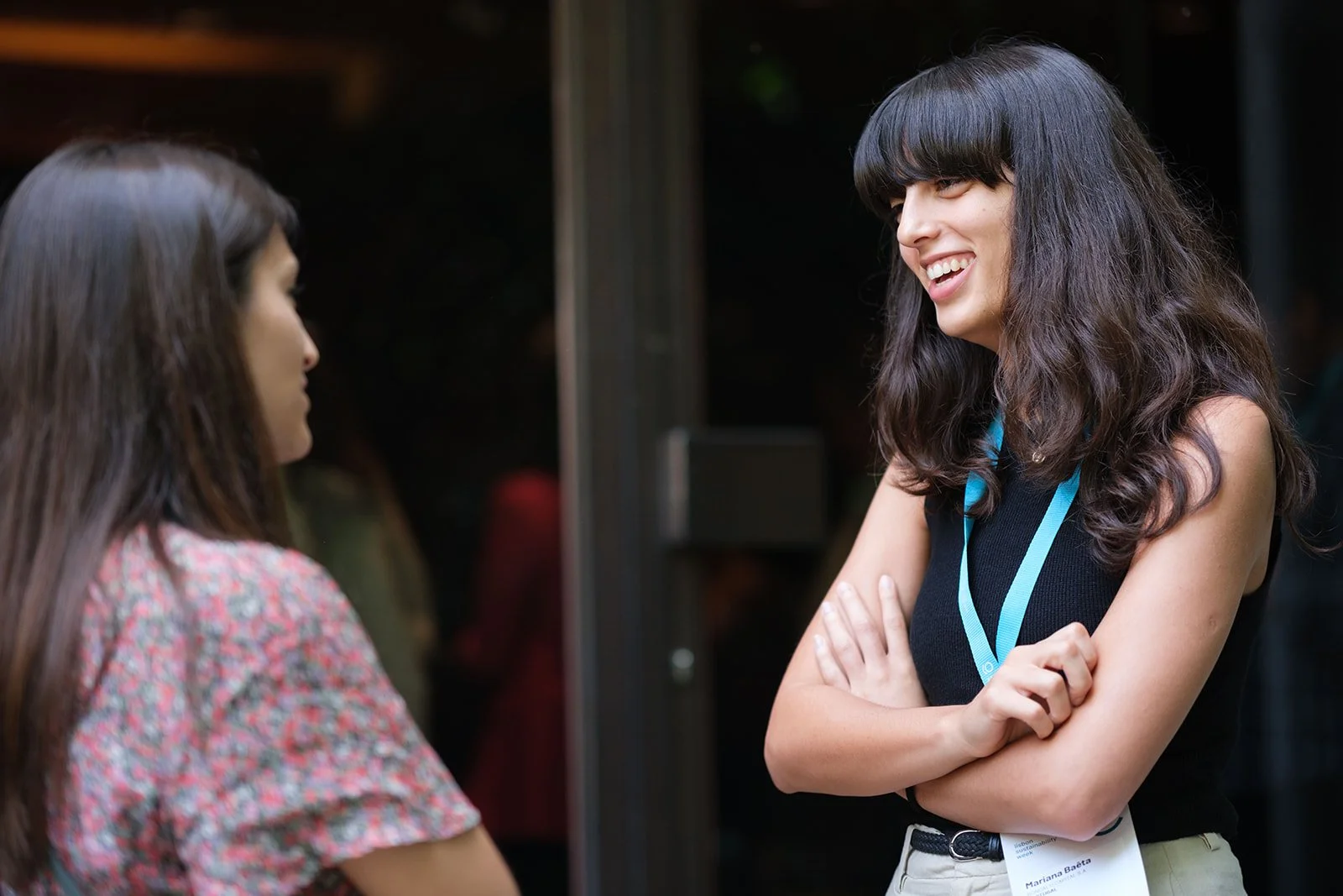 Two women engaged in conversation, one smiling with arms crossed, wearing a black sleeveless top and a conference badge, in an indoor or outdoor setting.