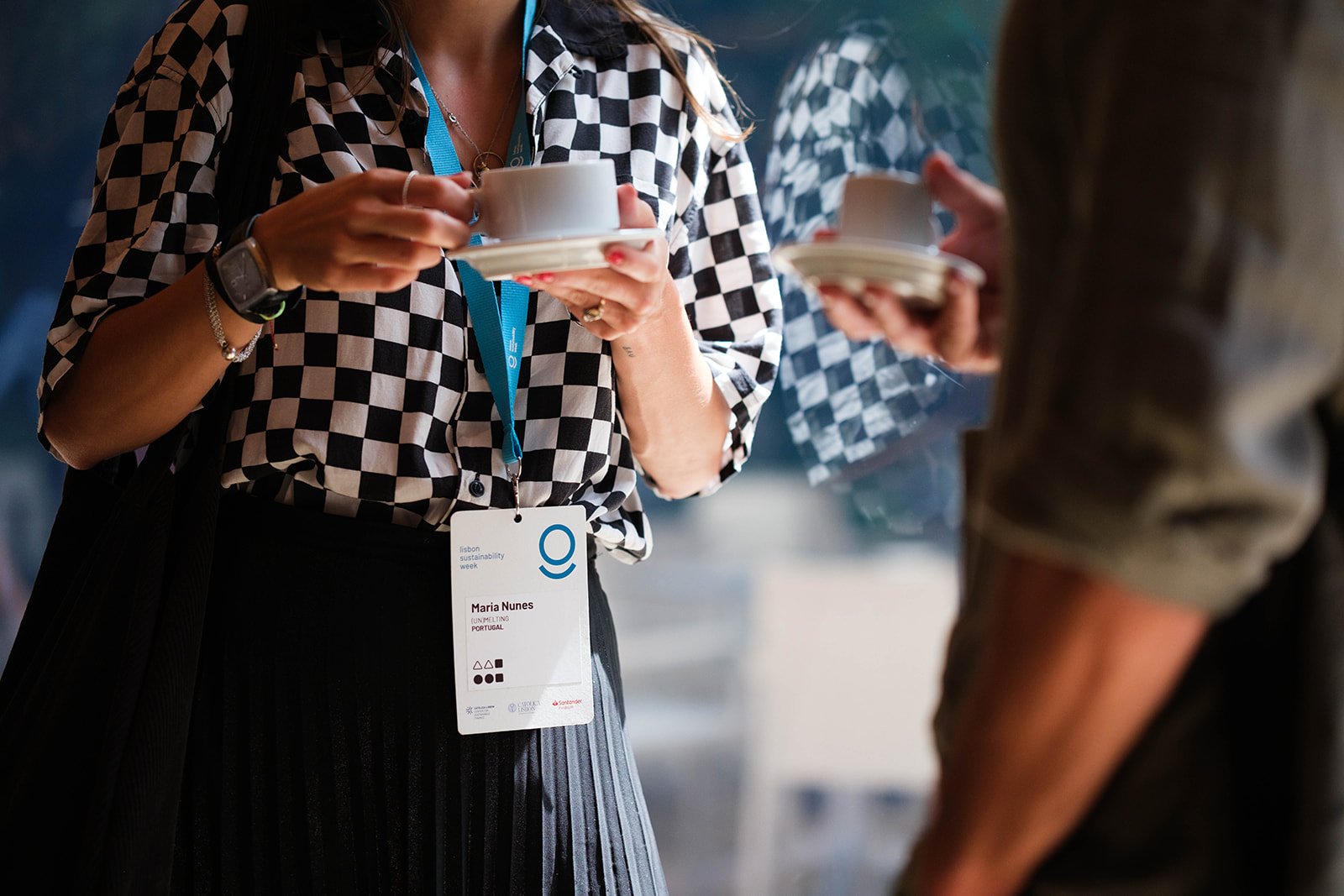 Two women holding coffee cups during a conversation at a conference or event, one of whom has a name tag that reads Maria Nunes from Portugal.