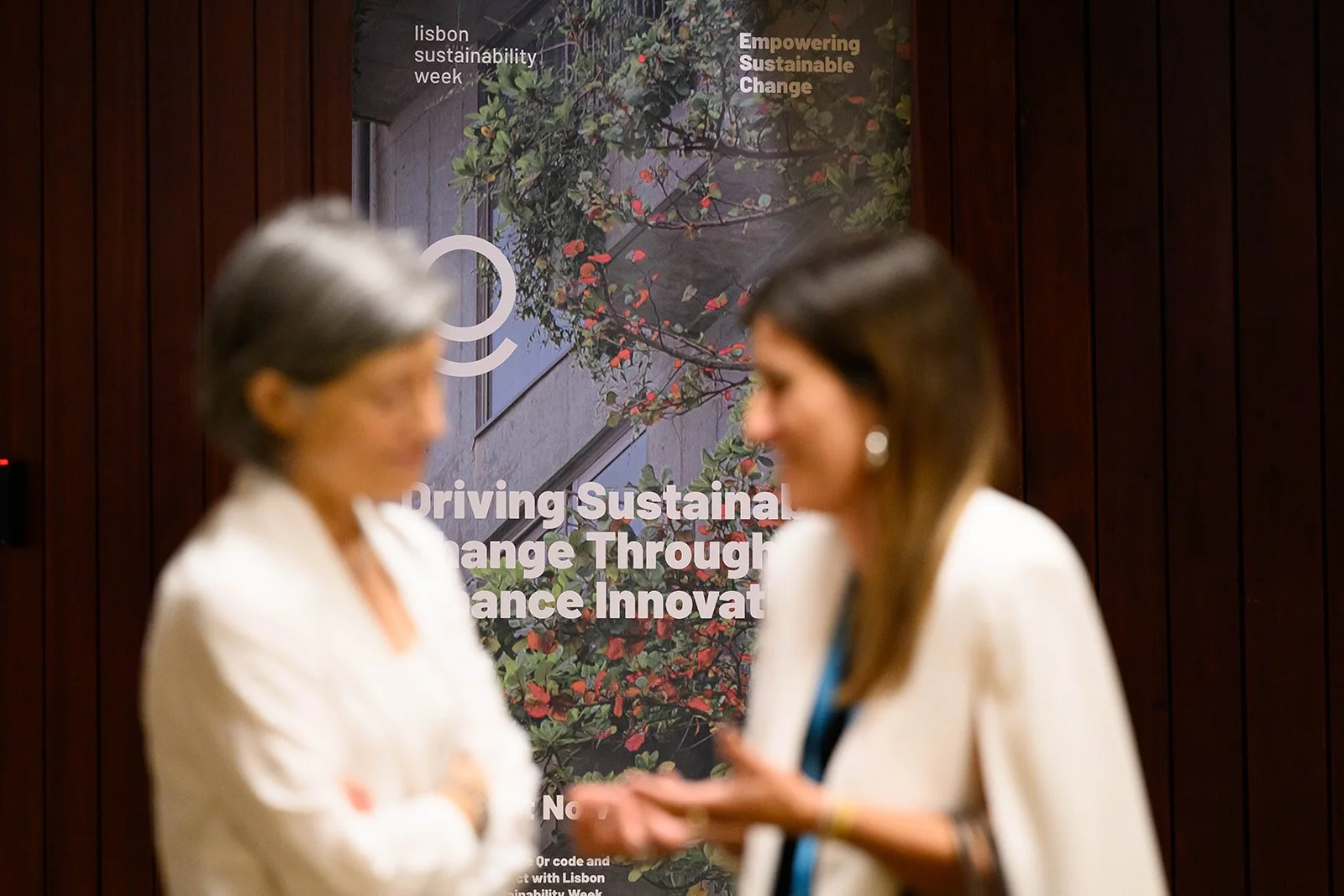Two women talking in front of a Lisbon sustainability week poster with text about driving sustainable change.
