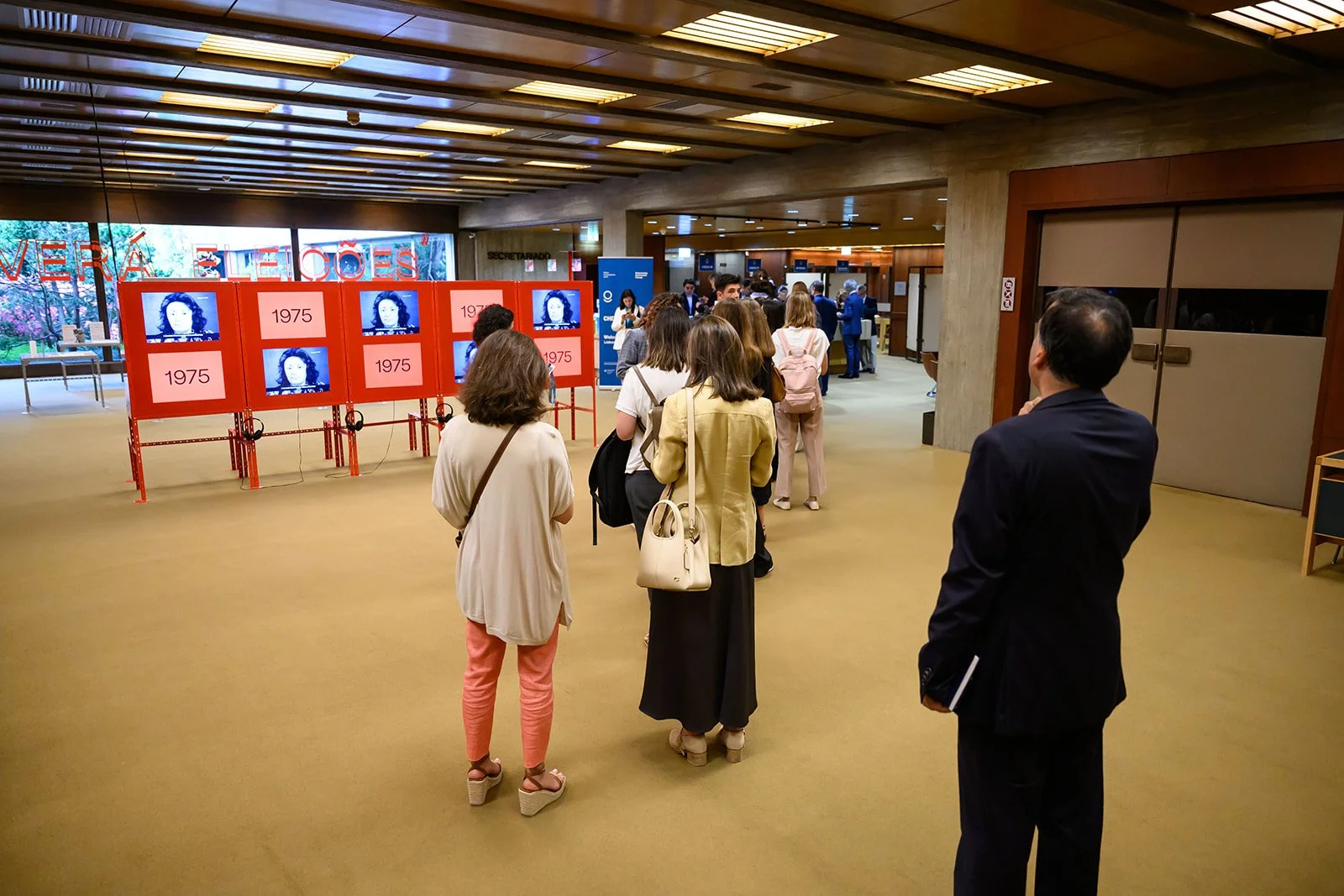 People standing in line in a gallery or museum, with red display boards showing images and the year 1975. Large windows in the background reveal trees and a suburban scene.