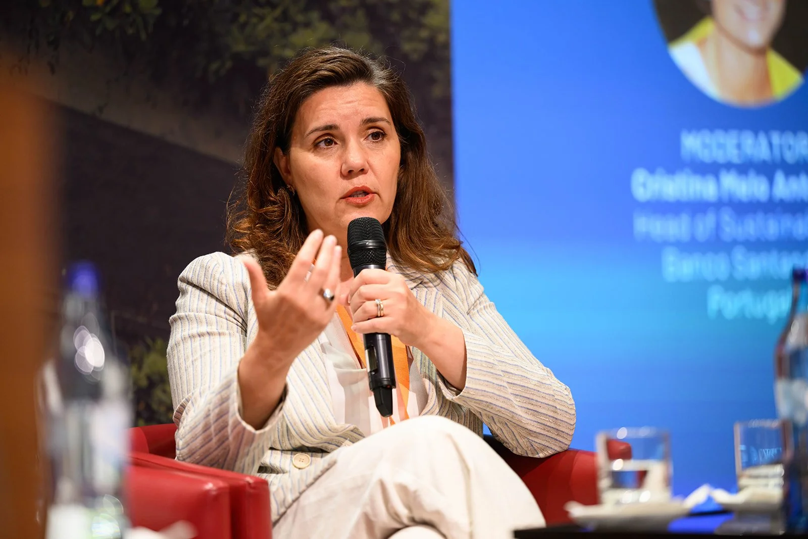 A woman speaking into a microphone during a conference or panel discussion, seated on a red chair with a blue presentation screen in the background displaying her name and title.