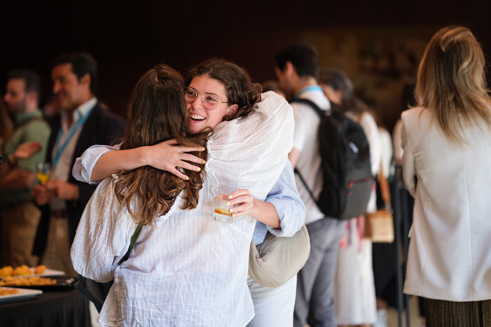 Two women hugging at a networking event or conference, with drinks in hand and other attendees socializing in the background.