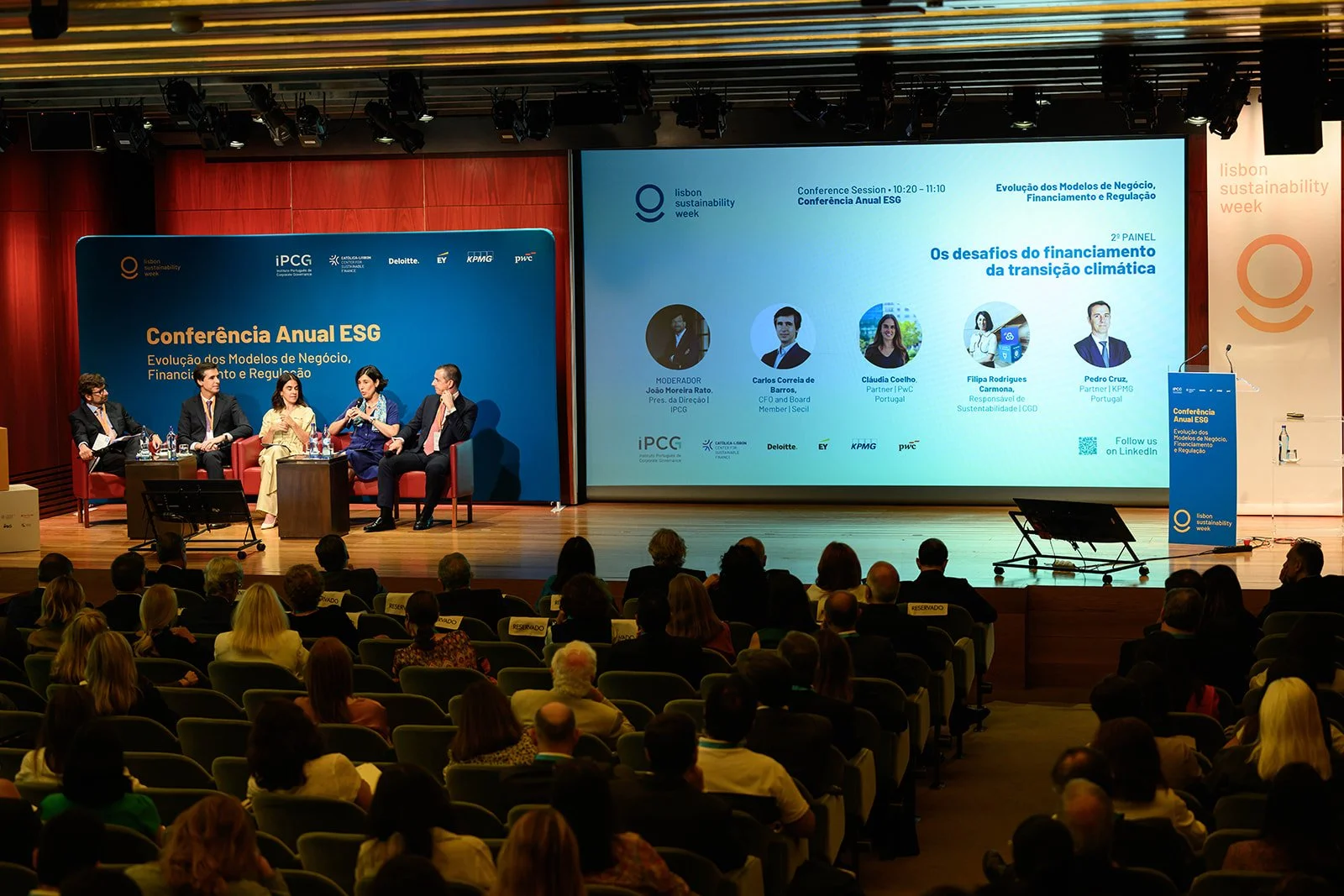 Panel discussion at a conference on sustainability week, with five speakers seated on stage, a large screen displaying session details, and an audience in attendance.