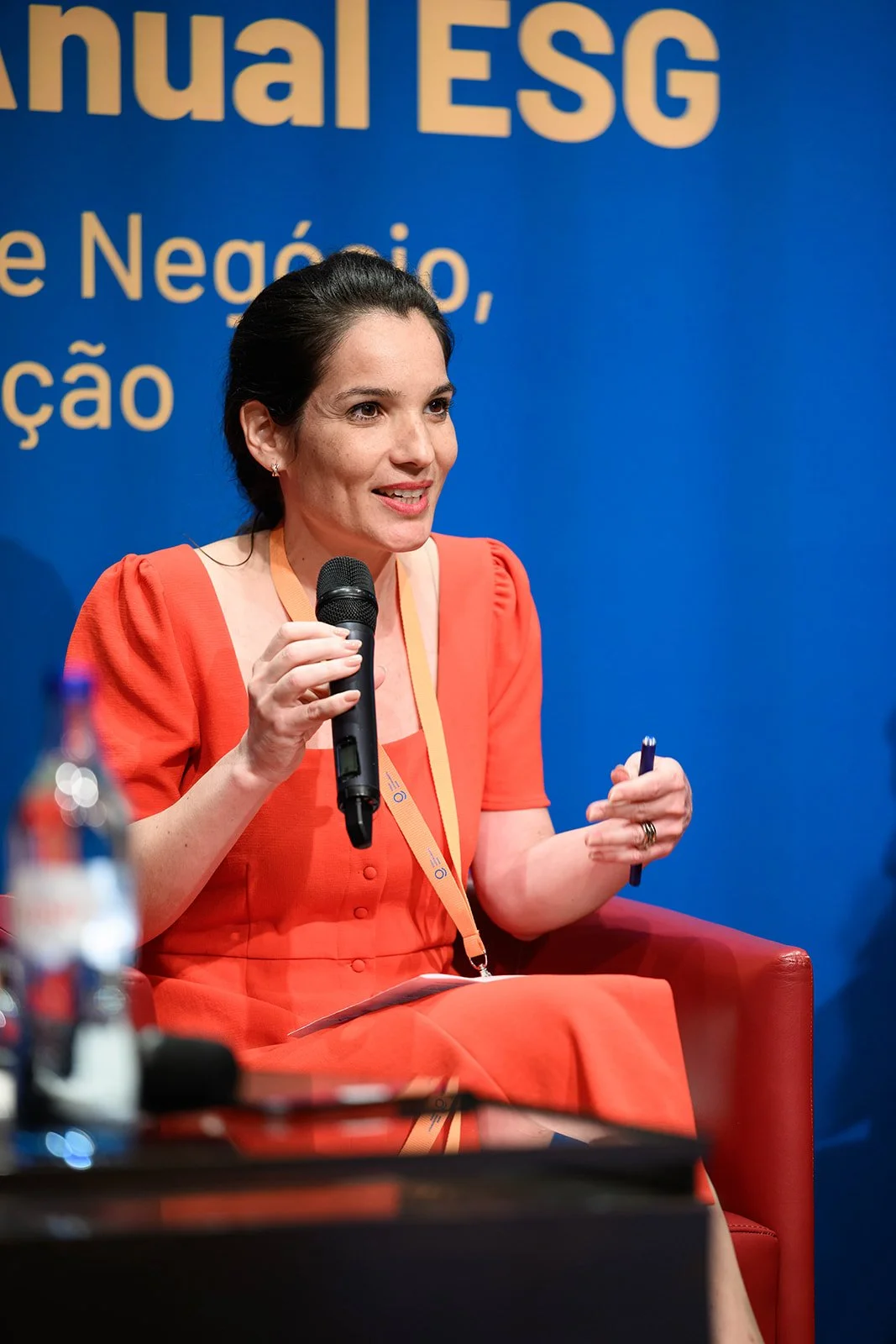 A woman in a coral red dress speaking into a microphone at an indoor event, with a blue backdrop that reads 'Annual ESG' and partial additional text.