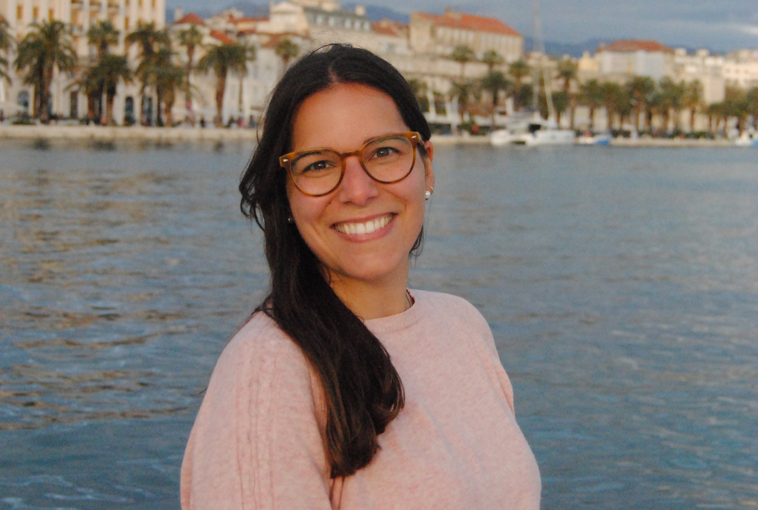 A woman with glasses and long dark hair smiling near a body of water with boats and buildings in the background.