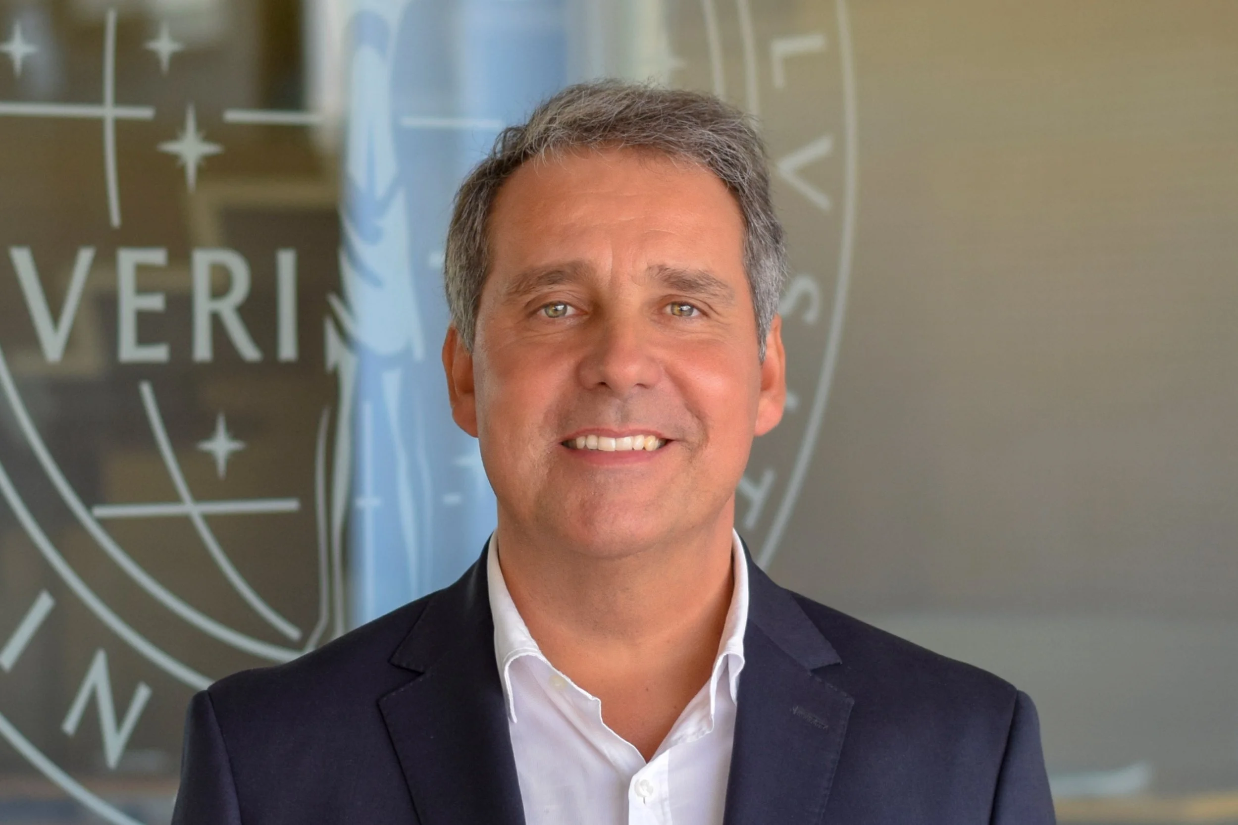 Smiling man in a suit jacket and white shirt standing in front of a blurred background with text.
