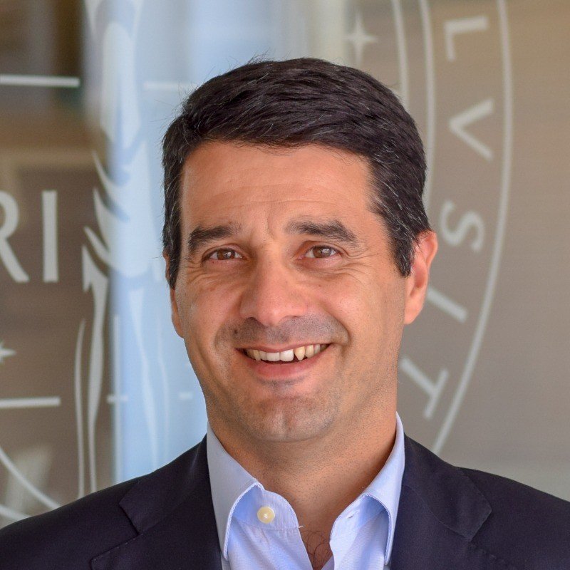 Smiling man in a suit with a blurred logo background.