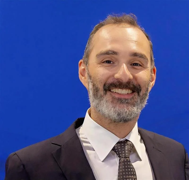 Man in a suit and tie smiling in front of a blue background.