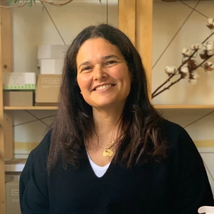 Smiling person with long hair wearing a black sweater standing in front of shelves with boxes and a decorative branch.