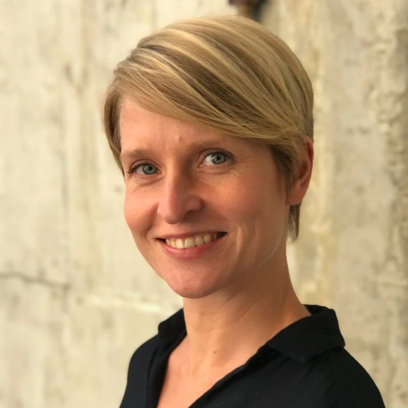Smiling person with short blonde hair standing against a textured wall.