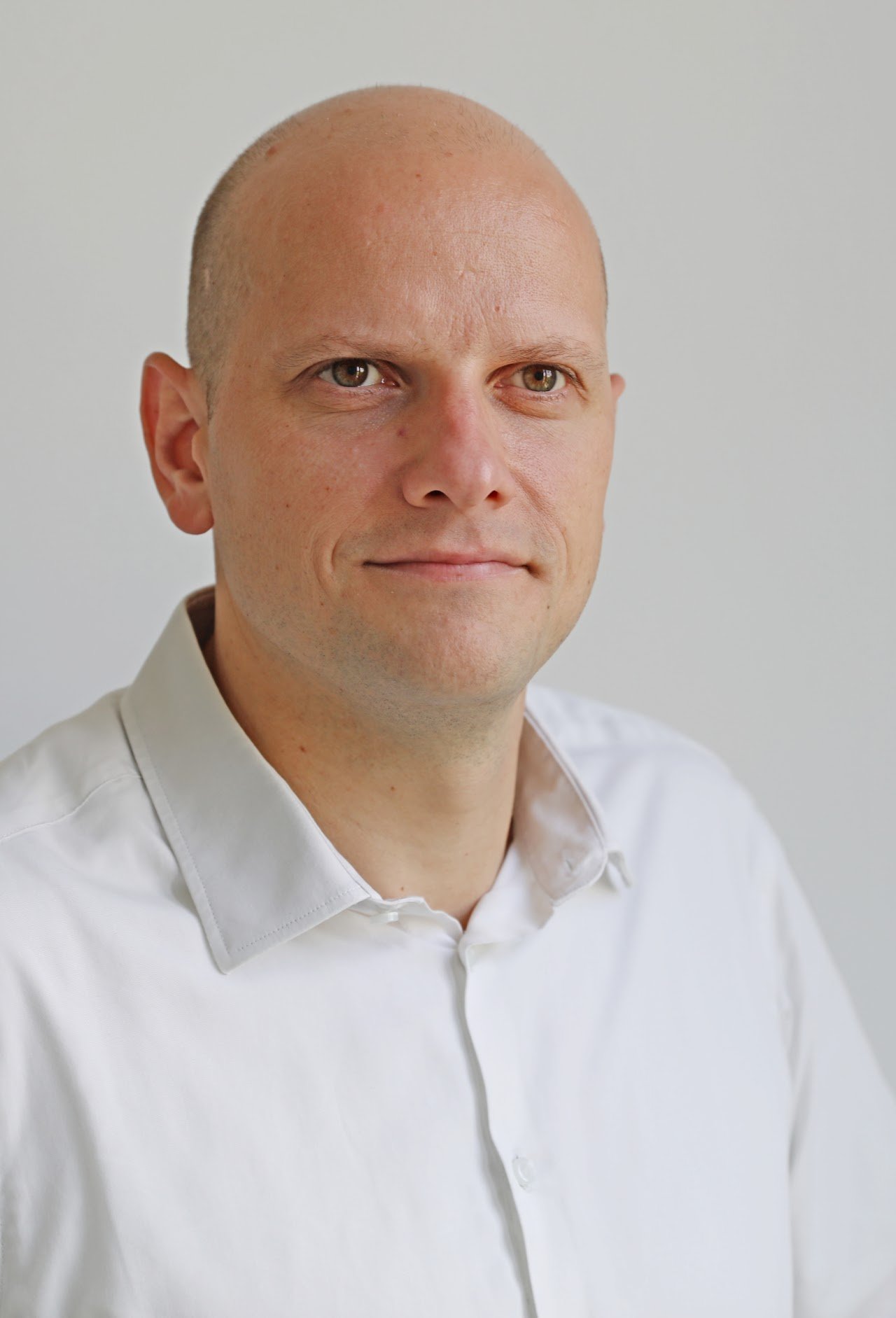 Close-up portrait of a man with a shaved head wearing a white shirt, looking at the camera with a slight smile against a plain white background.