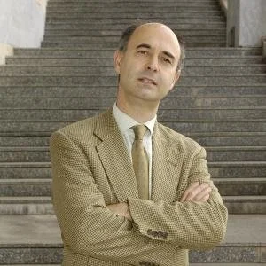 Person in a light brown suit standing with crossed arms in front of stone steps.