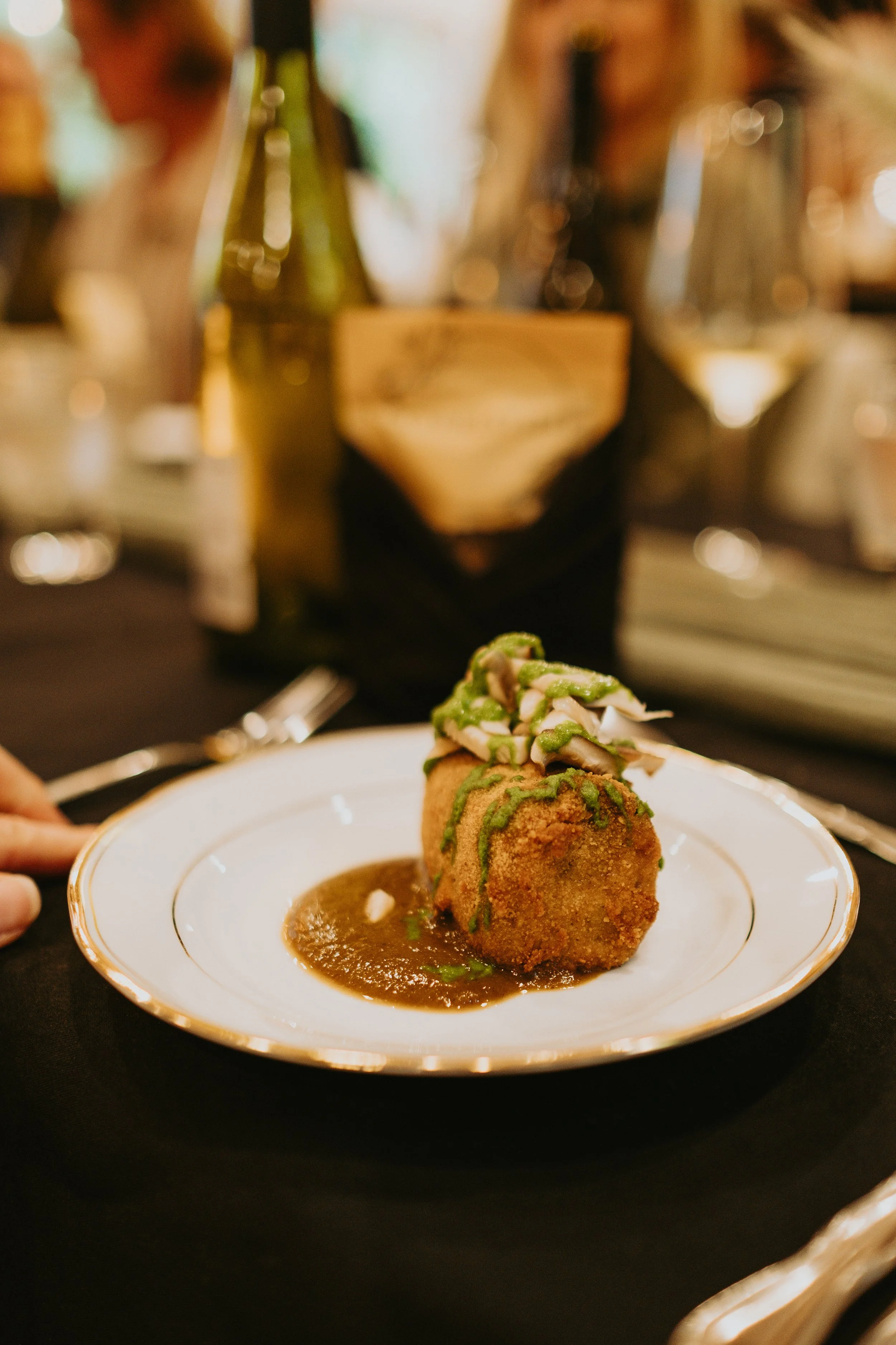 Un plat de croquettes de pain de viande garnies de sauce et de crème, servi avec une sauce brunâtre sur une assiette blanche avec une fine bordure dorée, dans un restaurant. En arrière-plan, une bouteille de vin, un verre de vin blanc, et une personne floue.