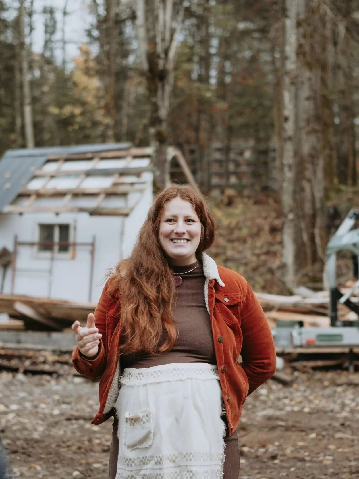Femme souriante debout dans une forêt, portant un tablier blanc et une veste marron avec un fond de cabanes en bois dans des arbres.