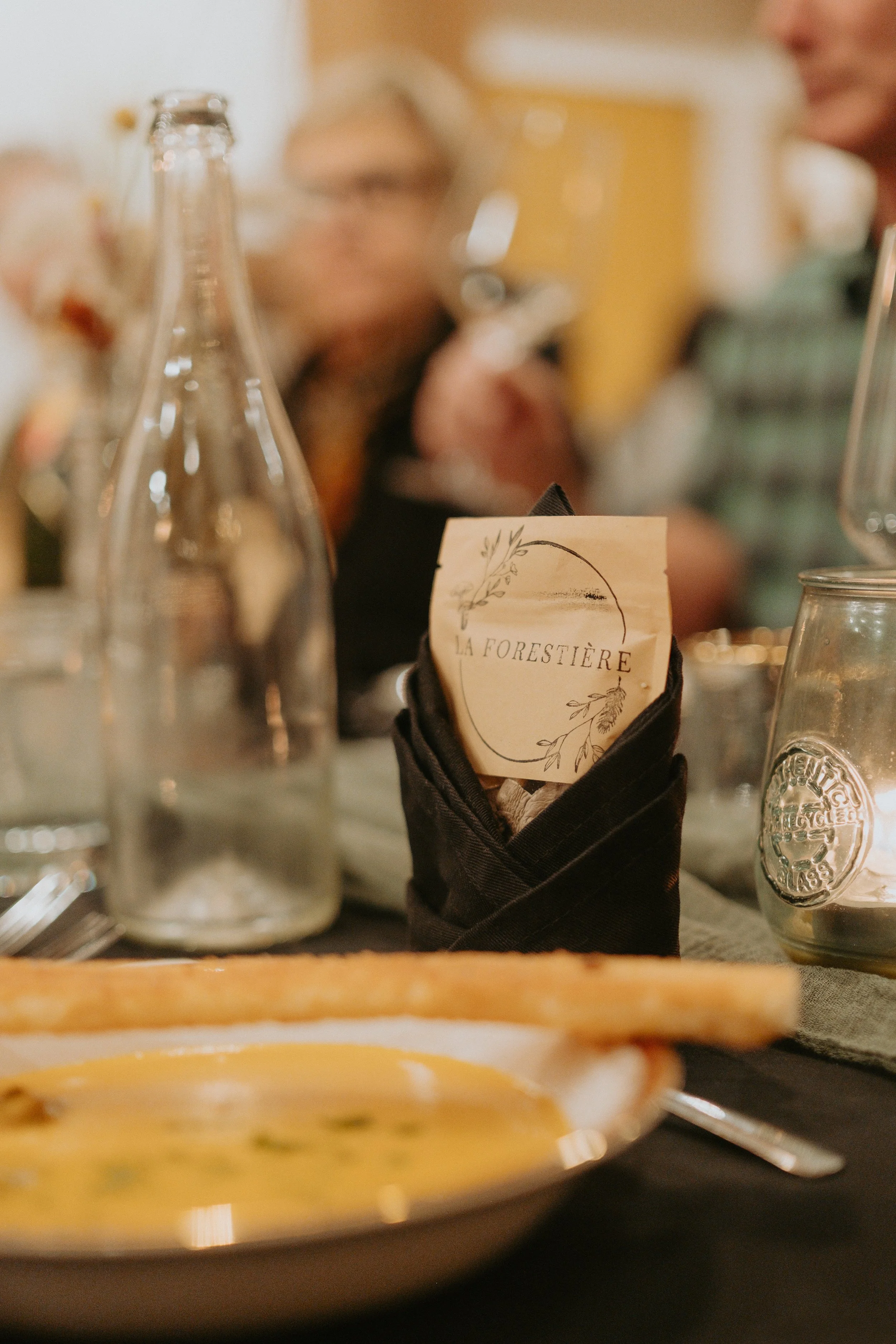 Une nappe de soirée avec une assiette de soupe à la courge, une bouteille en verre, un verre à vin, et une carte de menu portant le nom 'La Forestière' dans un porte-menu noir.