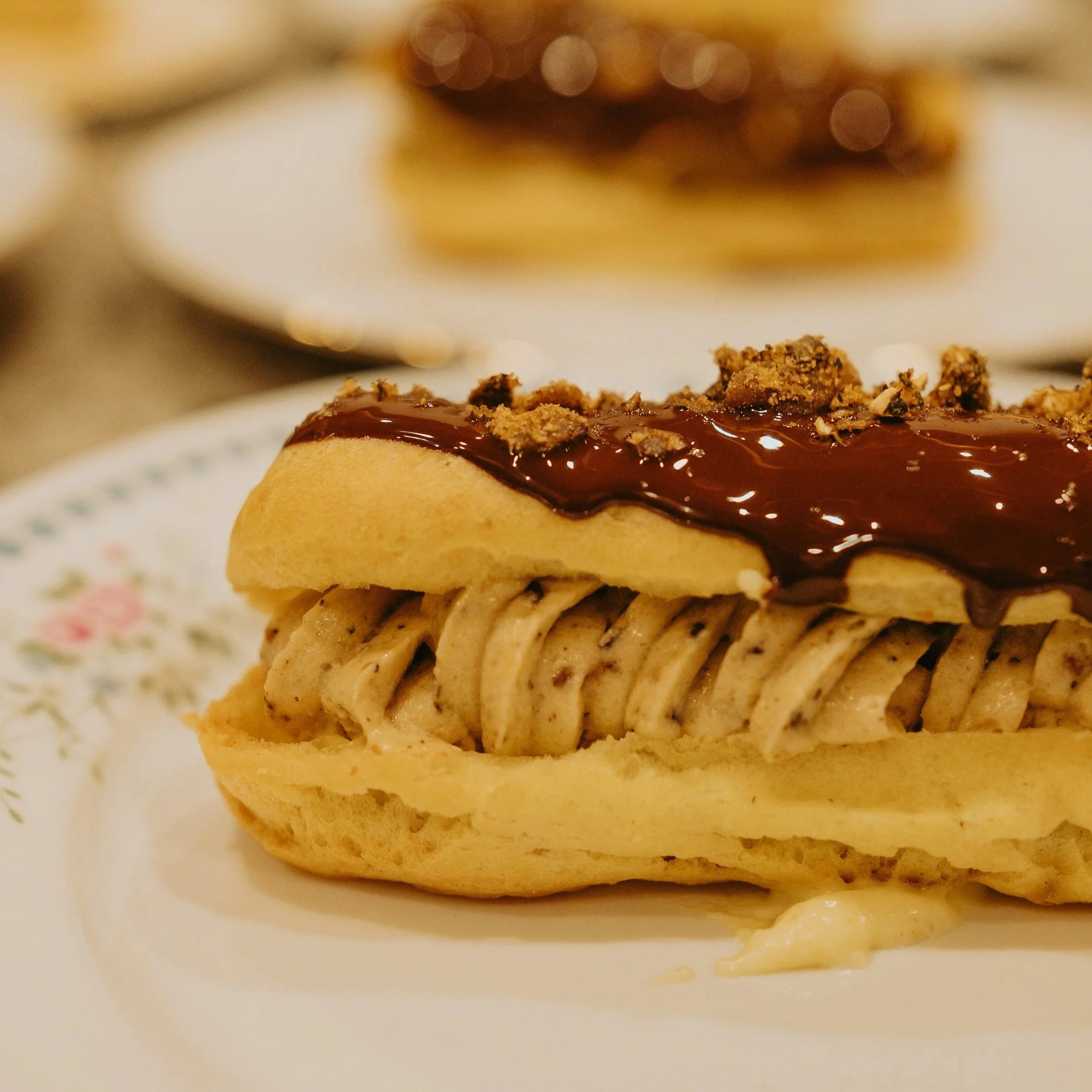 Une pâtisserie à éclairs garnie de crème à la vanille, recouverte de chocolat fondu et de biscuits croquants, servie sur une assiette blanche avec un motif floral.
