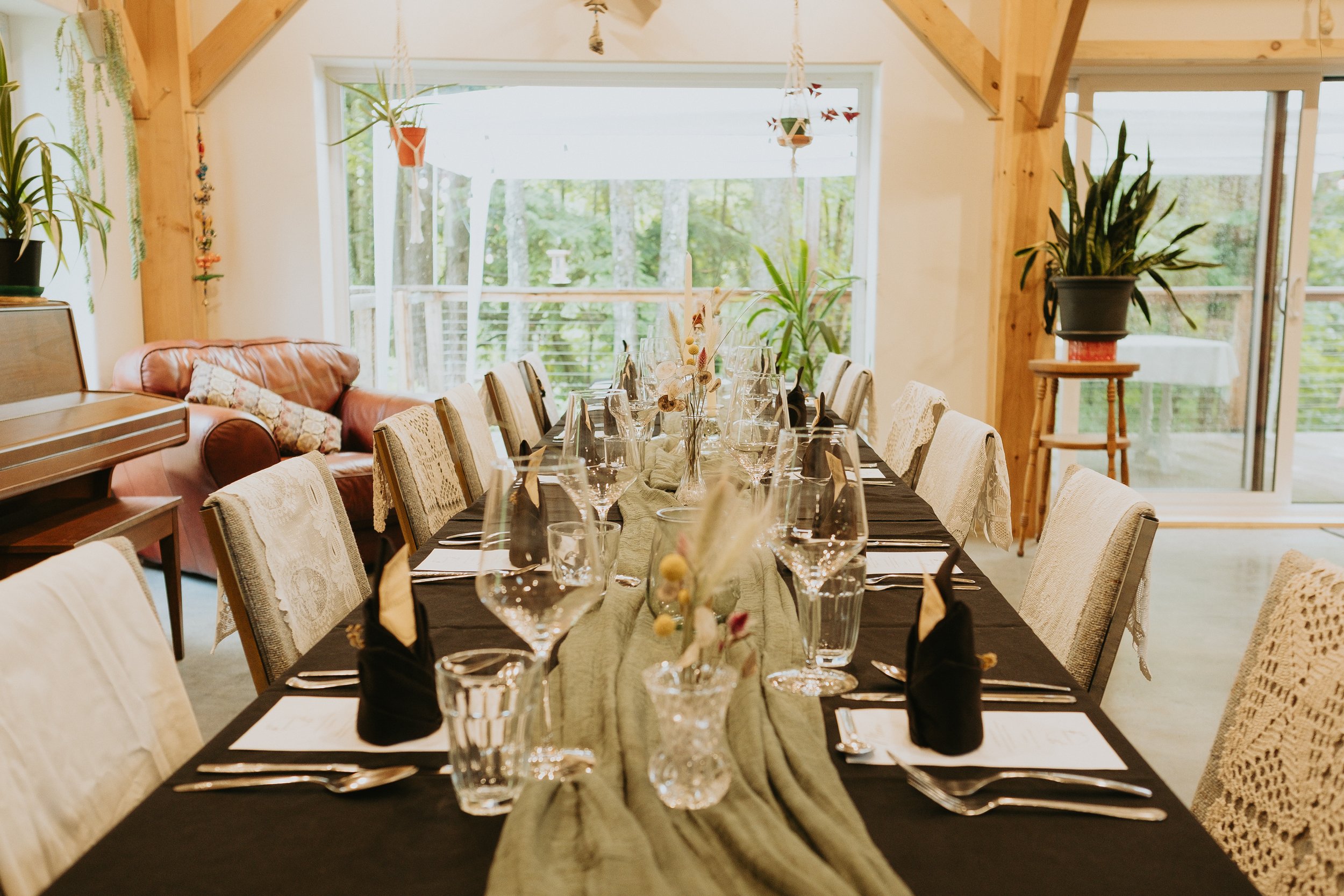Salle à manger avec une longue table décorée, entourée de chaises, dans un espace lumineux avec grande fenêtre extérieure, plantes et décoration intérieure