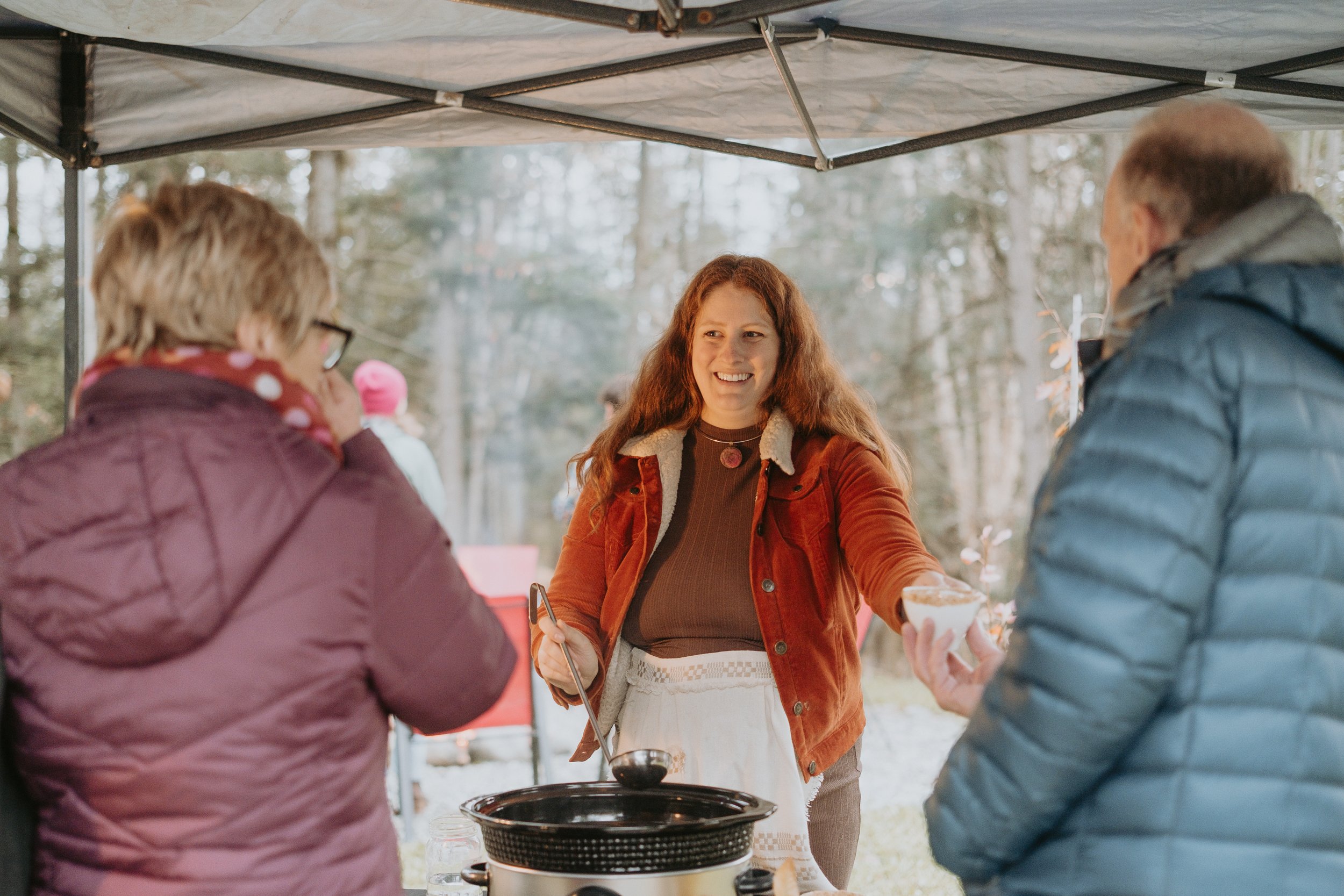 Trois personnes dans une fête en plein air, la femme au centre sert de la nourriture aux deux autres personnes sous une tente en forêt.