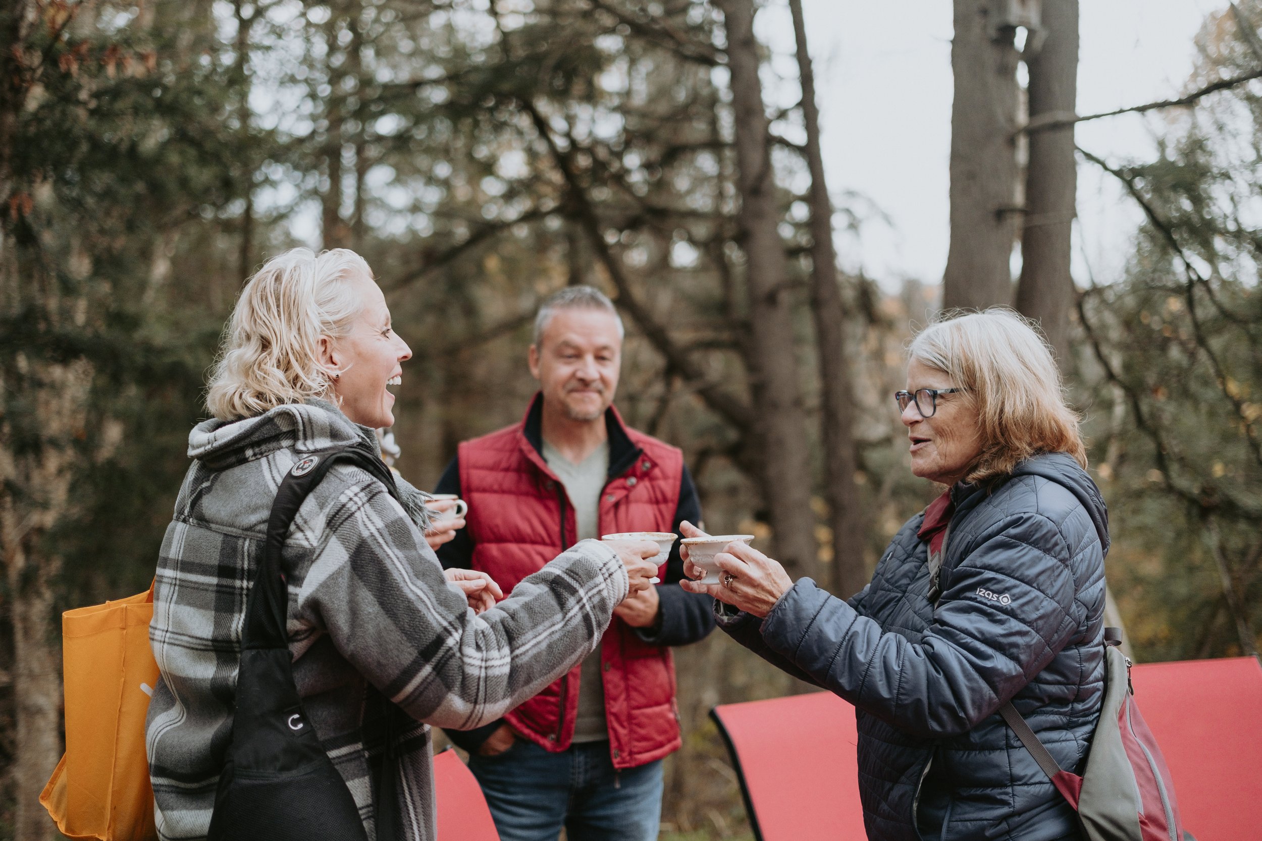 Trois personnes, deux femmes et un homme, sourient et jouent à un jeu de bâtons dans une forêt en automne, vêtus de vêtements chauds.