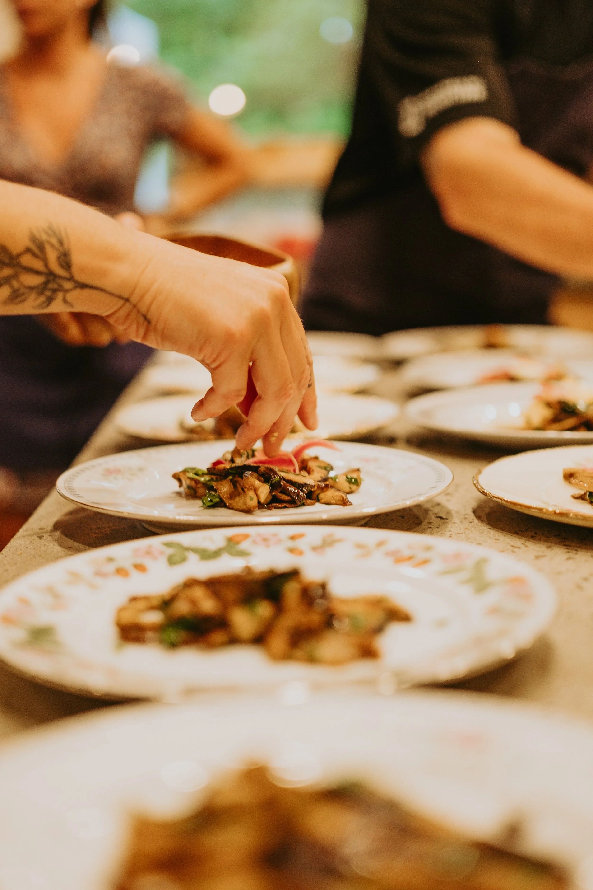 Main d'une personne en train de servir ou de disposer un plat de nourriture japonaise, avec plusieurs assiettes similaires sur la table.