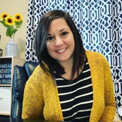 A young woman with dark hair and a smile, wearing a black and white striped shirt and a mustard yellow cardigan, sitting in a room with blue and white patterned curtains and a yellow sunflower arrangement in the background.
