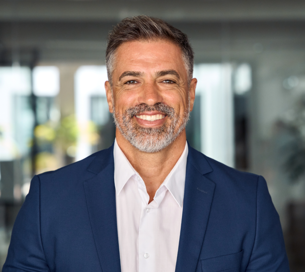 A smiling middle-aged man with gray hair and beard, wearing a navy blue suit and white shirt, standing in a modern, well-lit office building.