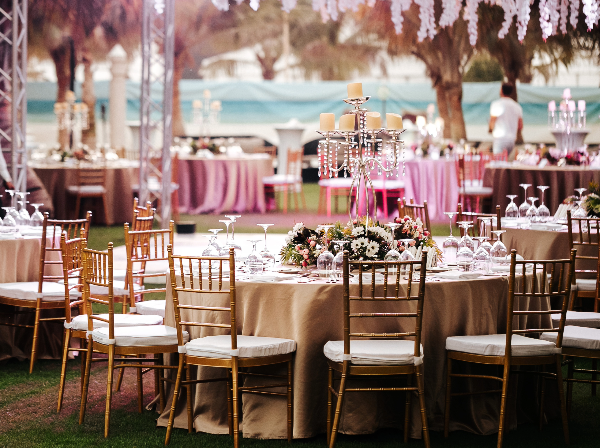 Elegant event setup with round tables covered in beige tablecloths, decorated with floral centerpieces, and surrounded by gold Chiavari chairs with white cushions.