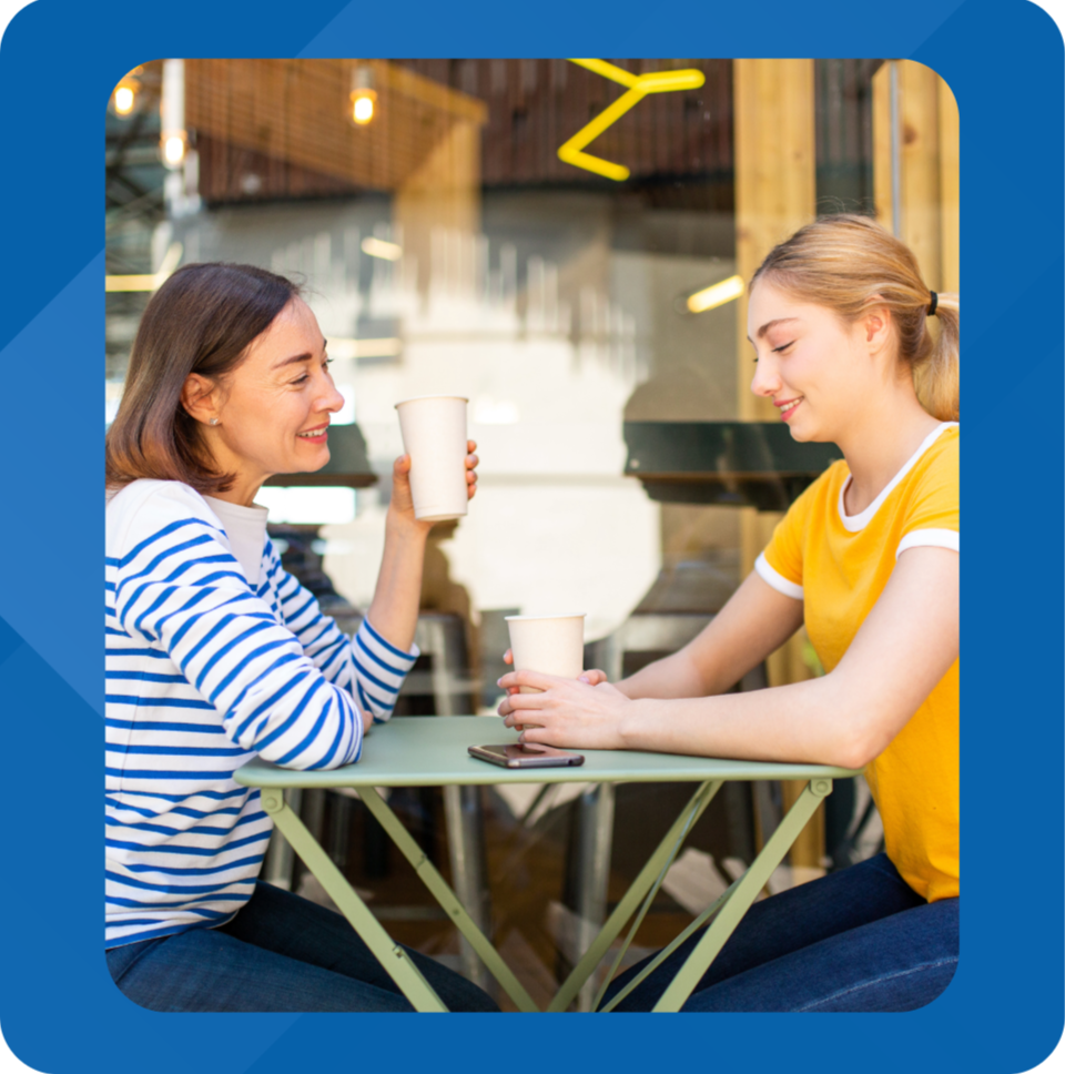 Two women sitting at a small table in a cafe, smiling and chatting while holding disposable coffee cups.