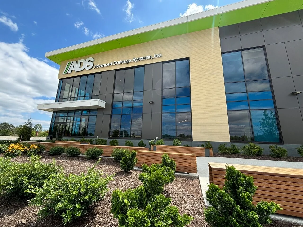 Modern building with large glass windows and a green accent on the roof, labeled 'ADS Advanced Drainage Systems, Inc.' with landscaped bushes and wooden benches in the foreground.