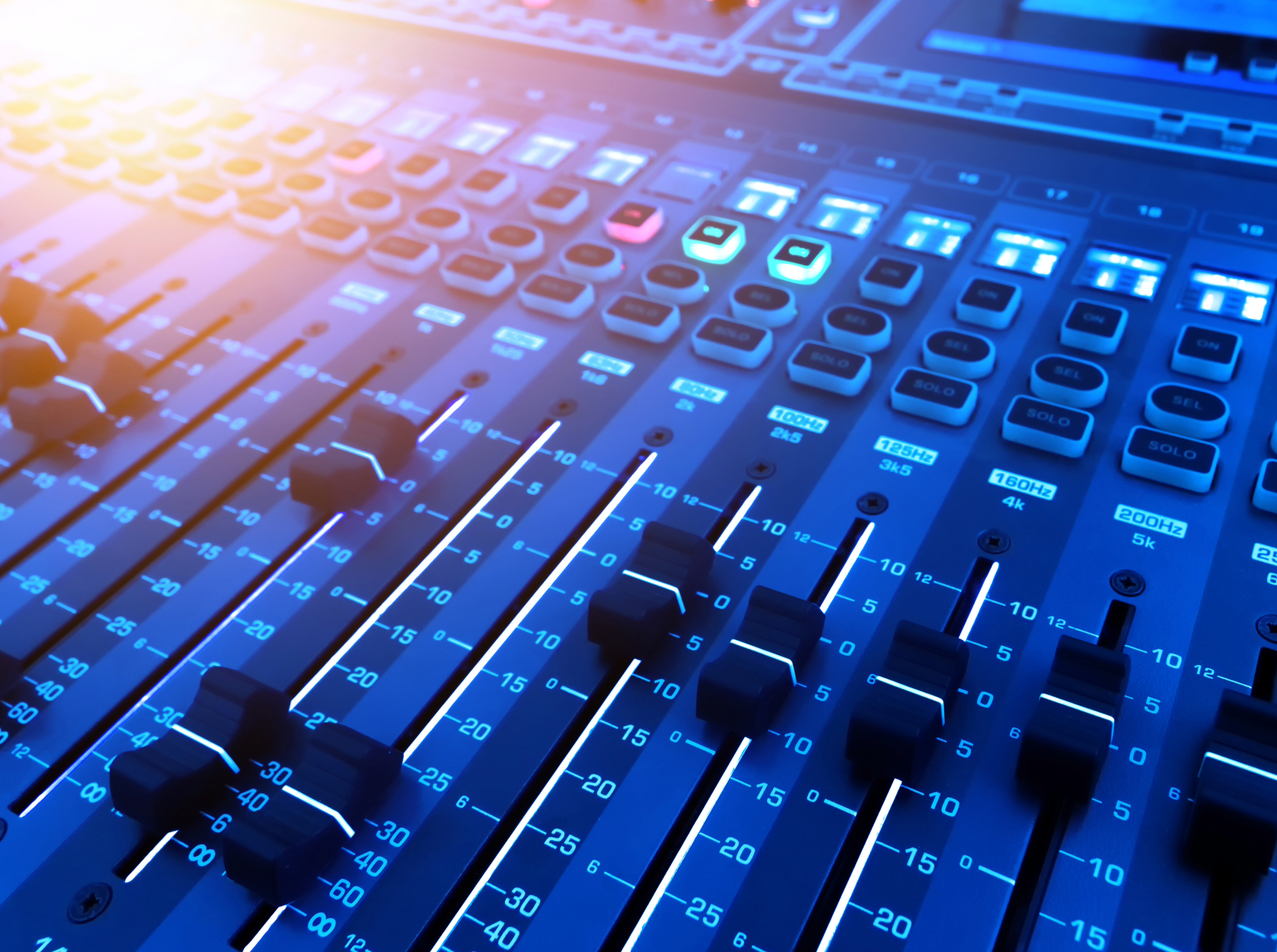 A close-up of a professional audio mixing console with illuminated faders, knobs, and digital displays.